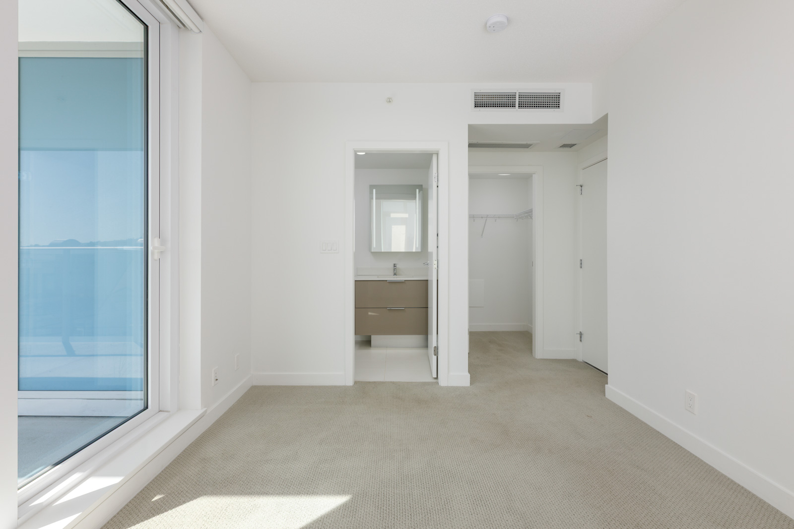 Empty, carpeted room with large window, white walls, an open door leading to a bathroom with a vanity, and another door opening to a closet.