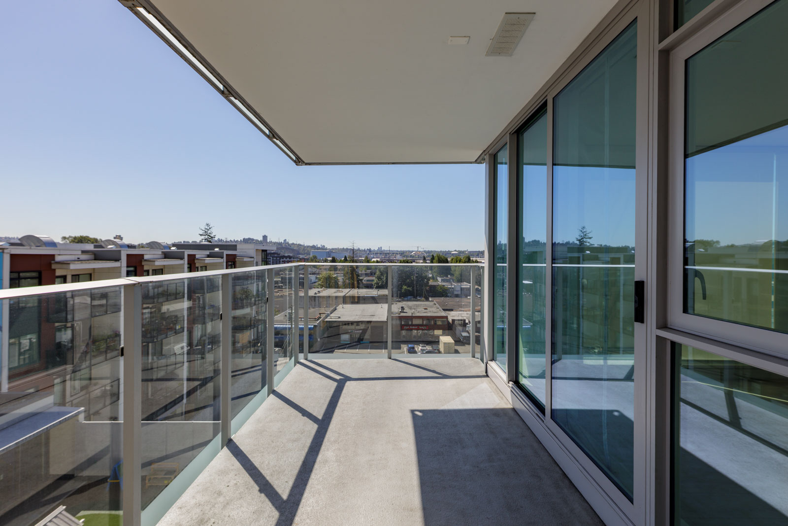Glass-walled balcony with a concrete floor attached to a modern building, overlooking rooftops and a distant city skyline under a clear blue sky.