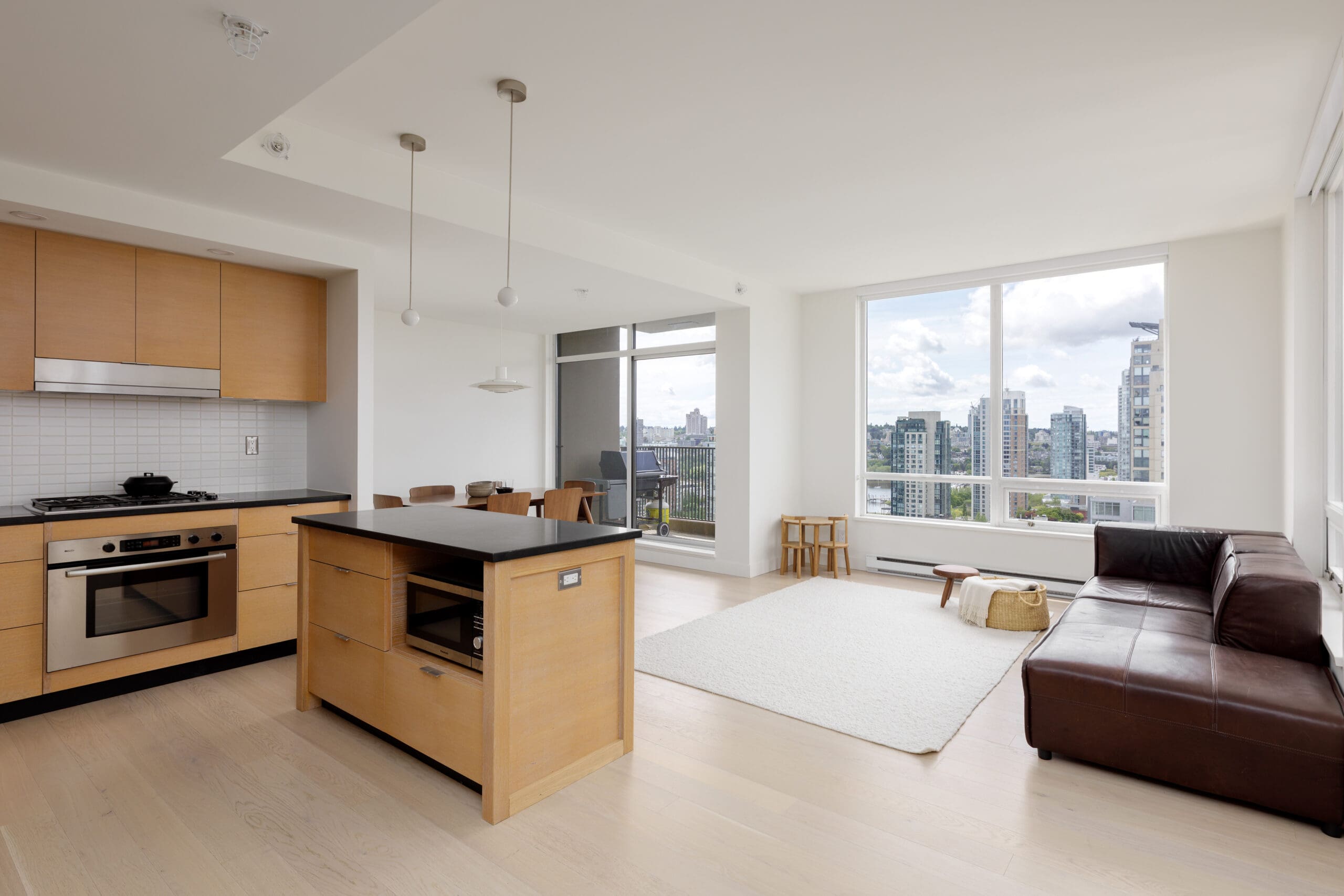Modern open-plan apartment with a kitchen, island, and living area featuring large windows, a brown leather sofa, and city views.