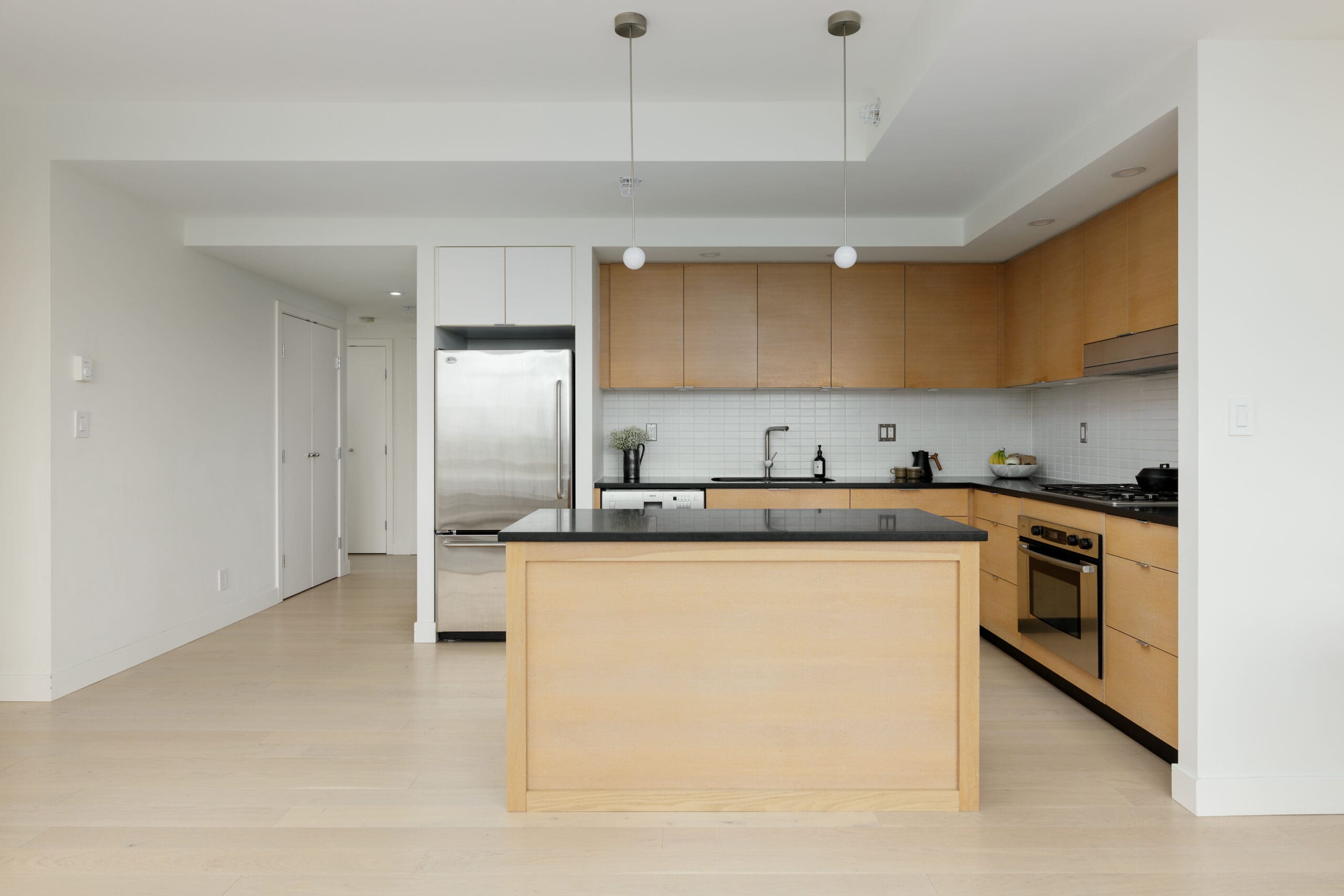 Modern kitchen with light wood cabinets, a central island, stainless steel appliances, and pendant lights, featuring a minimalist design and neutral color palette.