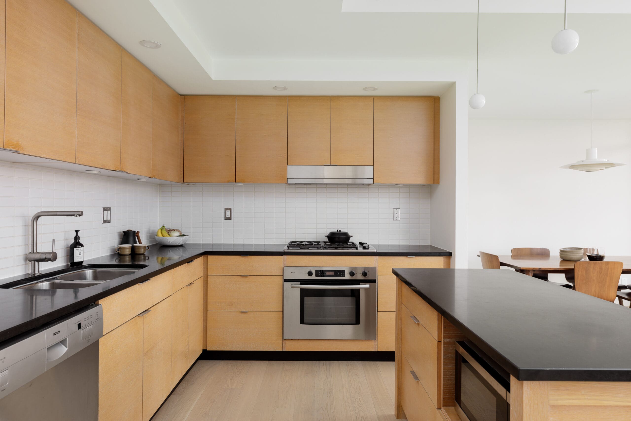Modern kitchen with light wood cabinets, black countertops, stainless steel appliances, and a dining area visible in the background.