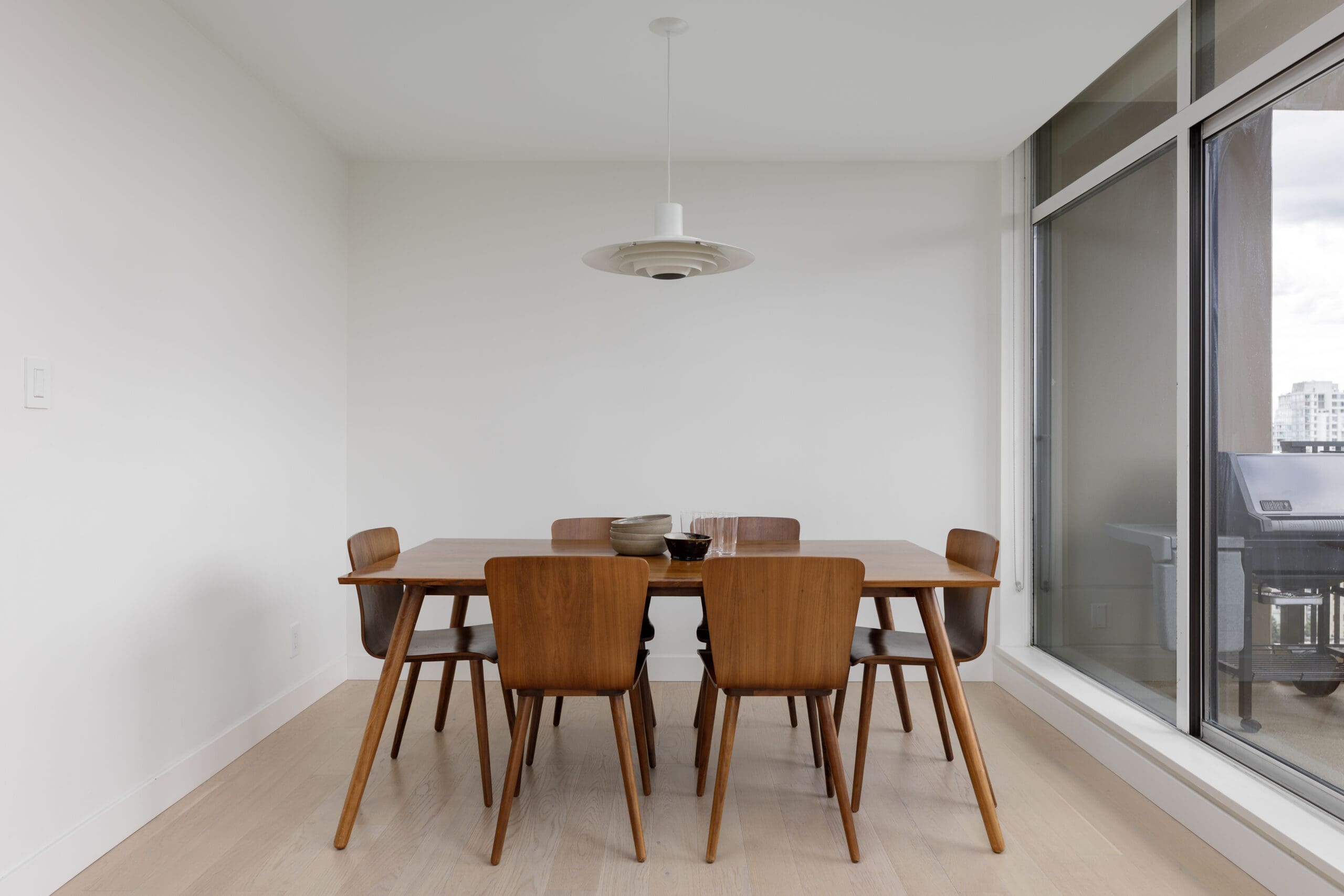 A modern dining area with a wooden table, six chairs, a pendant light above, and large windows showing a city view.