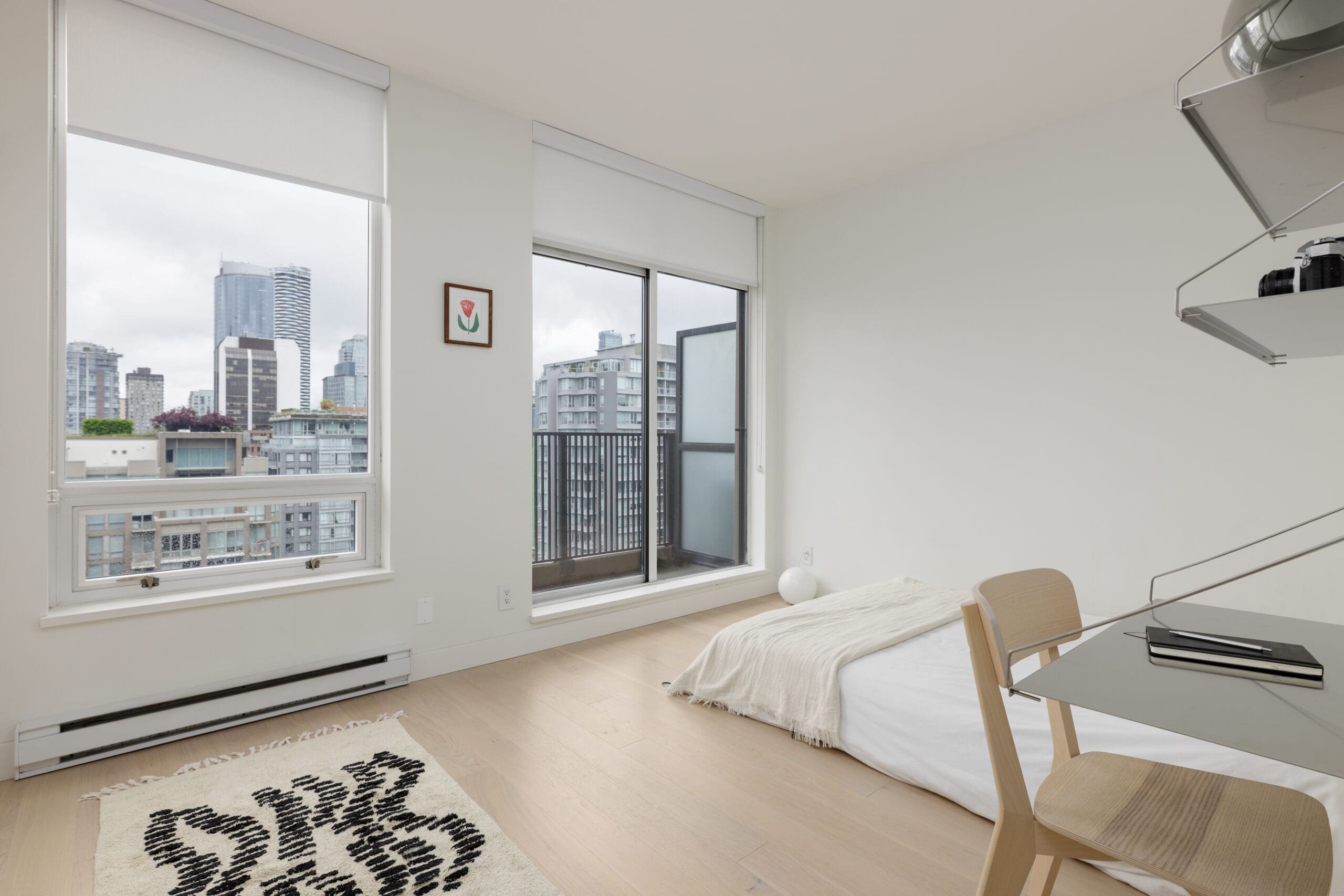 Minimalist bedroom with a low bed, desk, chair, and rug, featuring large windows and a sliding door that open to a city view with tall buildings.