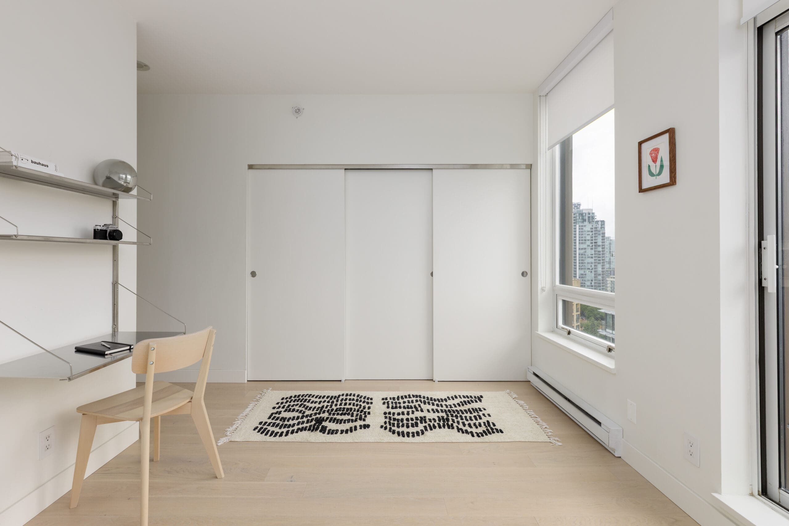 Minimalist room with light wood floors, a desk and chair on the left, a black and white rug, large closet doors, and a window with city buildings visible outside.