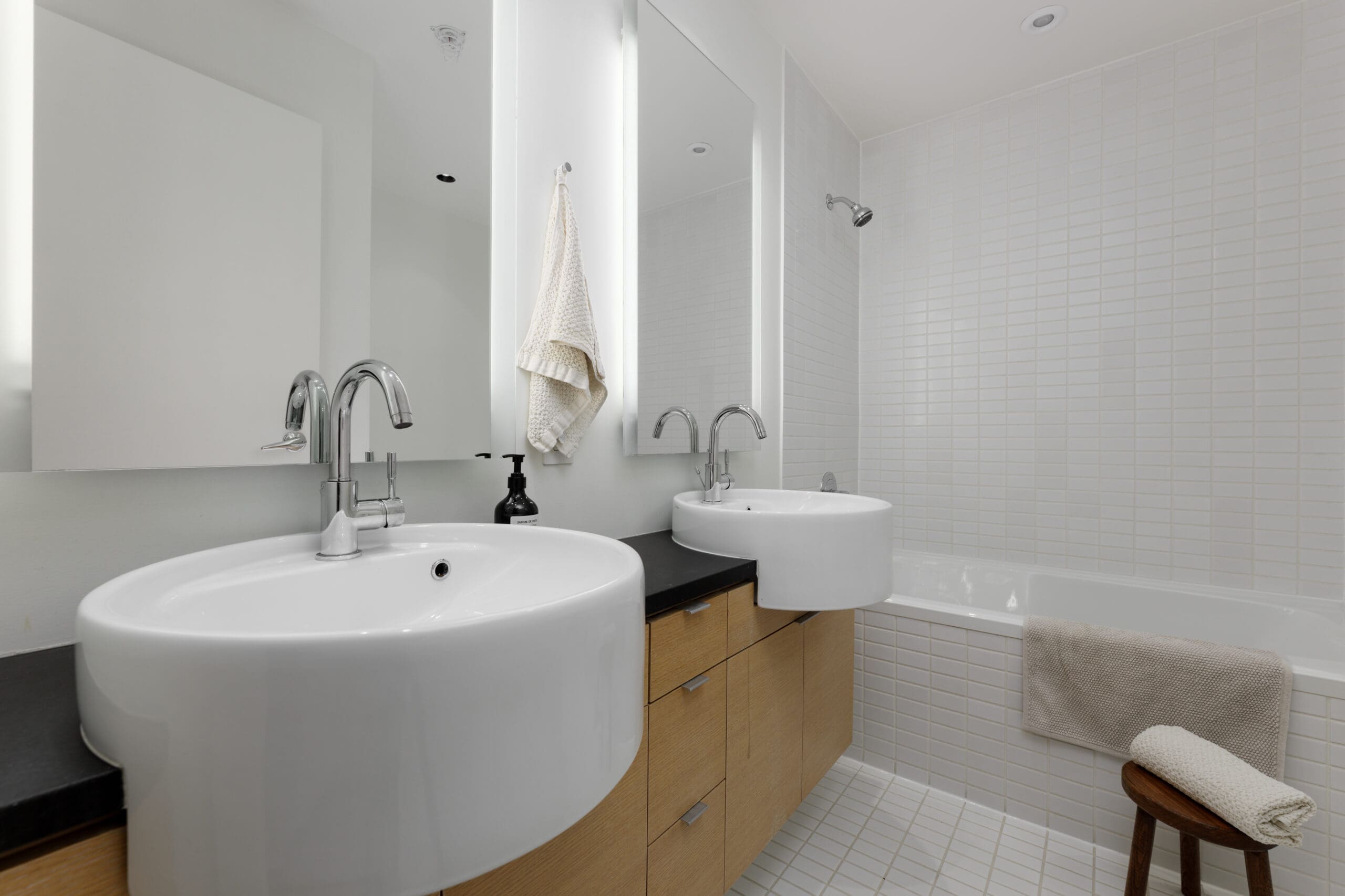 Modern bathroom with double sinks, wall-mounted mirrors, light wood cabinetry, a bathtub with a towel, and white tiled walls and floor.