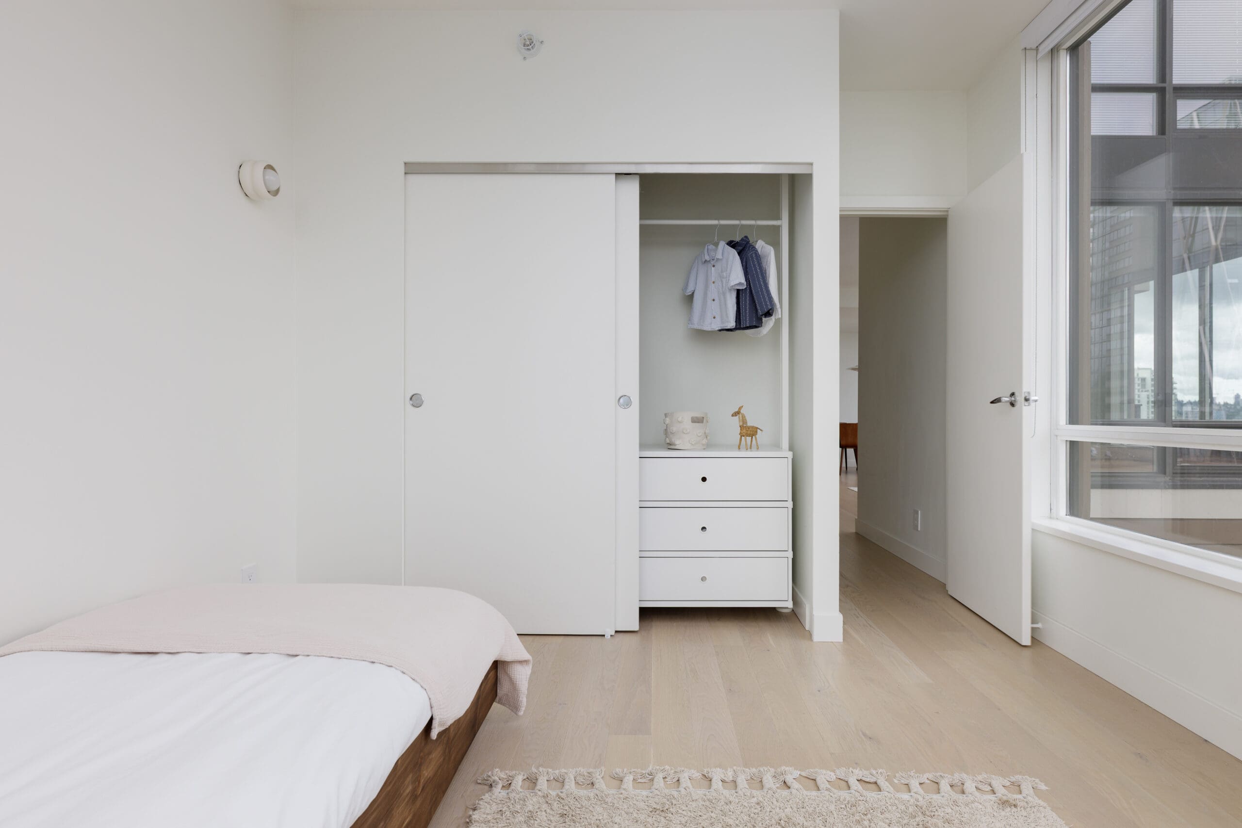 Minimalist bedroom with a bed, an open closet showing a few hanging clothes, drawers, and a window letting in natural light. The walls and décor are neutral in color.