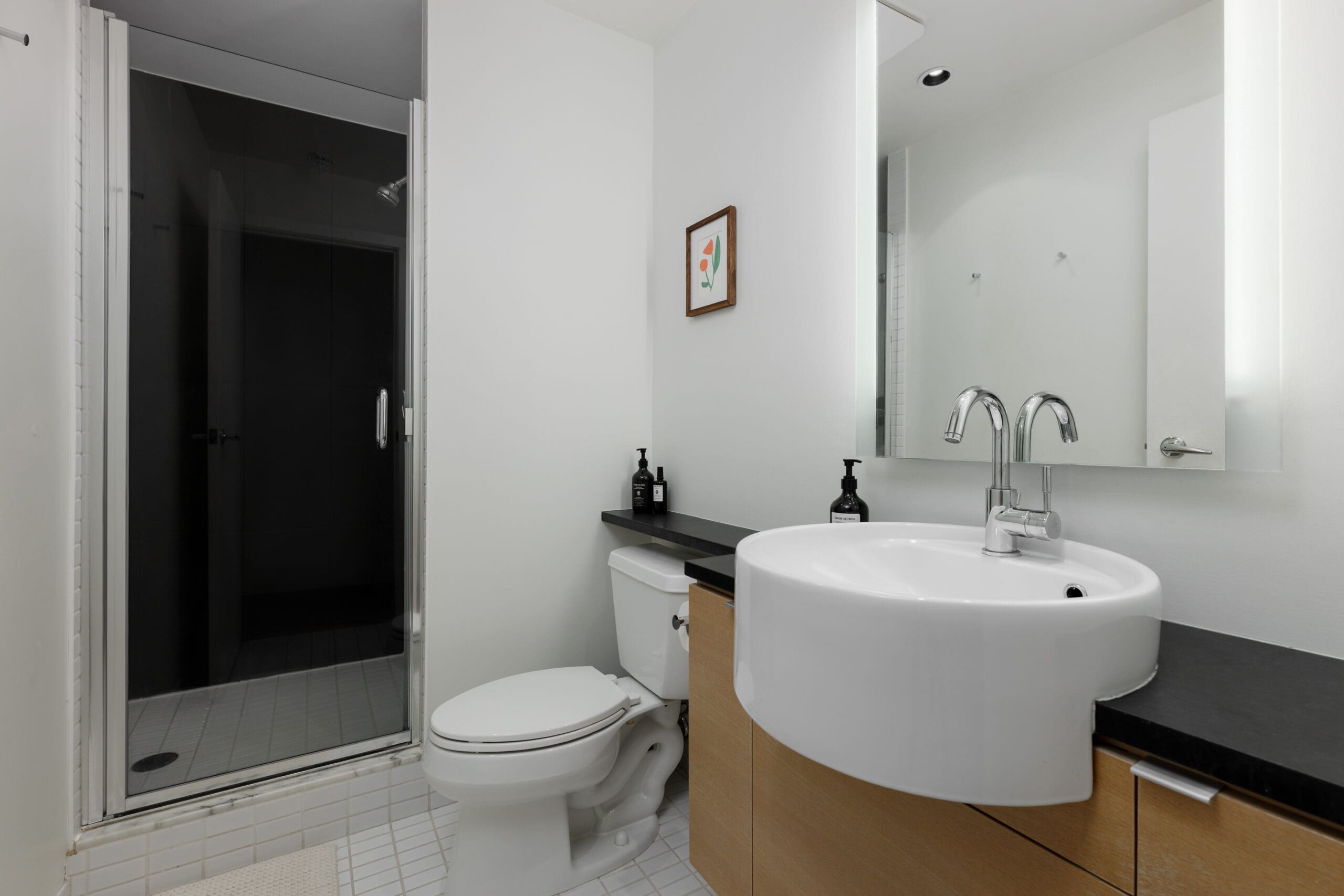 Modern bathroom with a glass-door shower, white toilet, round sink on a wooden vanity, wall-mounted mirror, and minimal decor.
