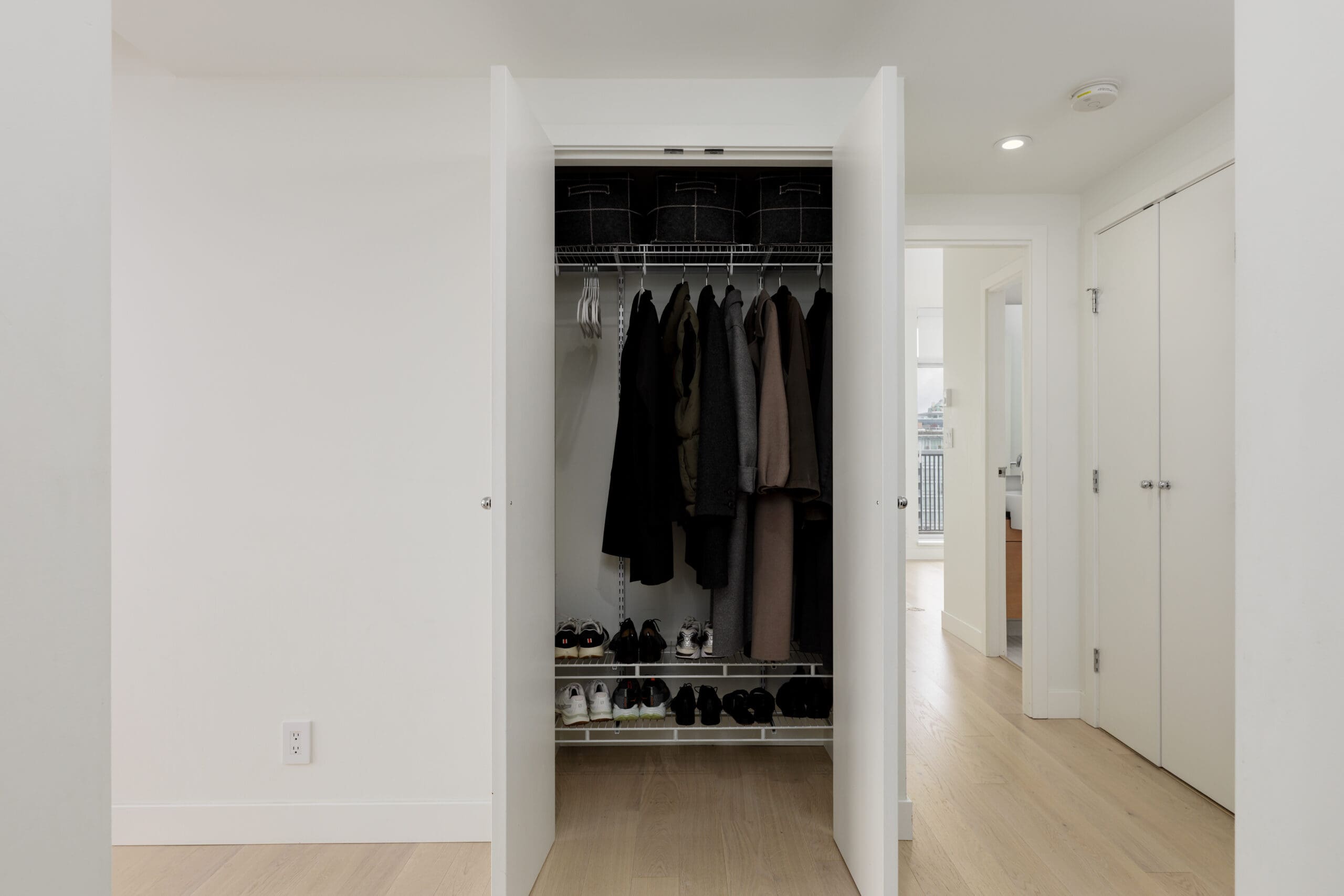 Open closet with coats hanging on a rack, shoes organized on shelves below, and storage boxes on the top shelf in a bright, minimalist room.