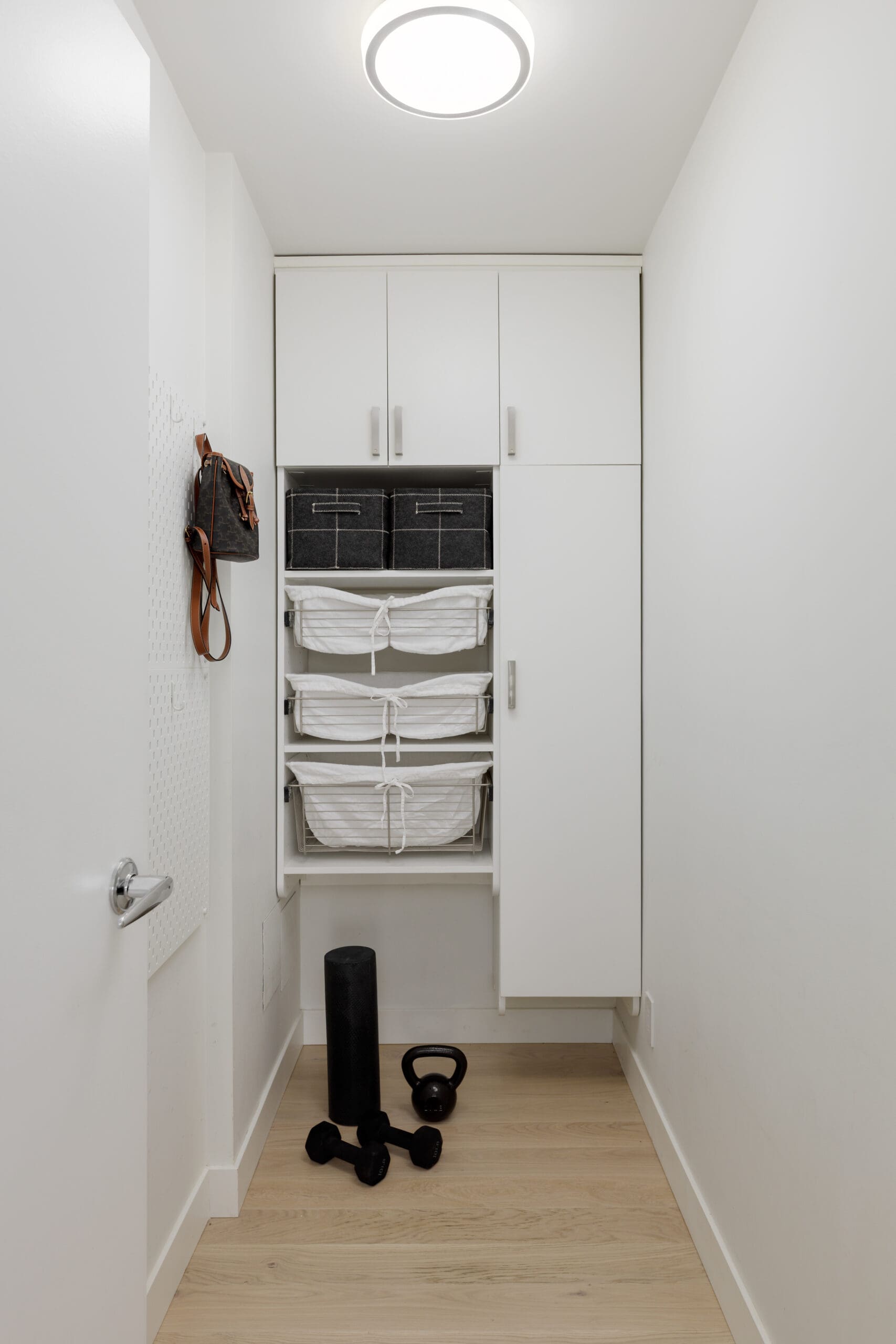 A small, white laundry room with cabinets, three white baskets, black storage bins, a black bag hanging on the wall, and exercise equipment on a wooden floor.
