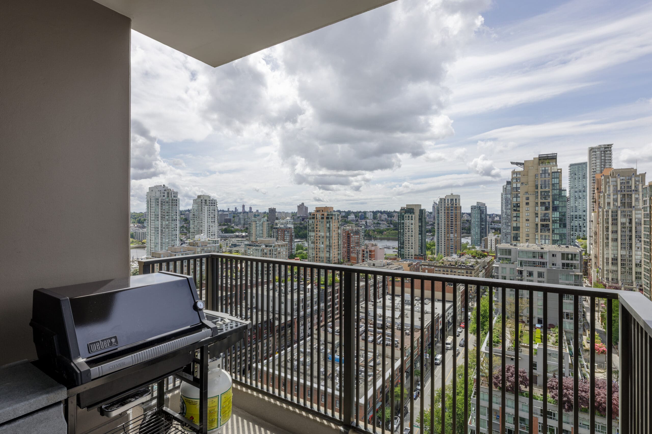 A balcony with a gas grill overlooks a cityscape featuring high-rise buildings and a partly cloudy sky.