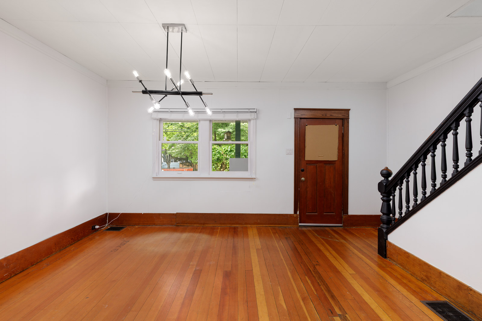 Empty room with hardwood floors, white walls, a window, wooden front door, black staircase railing, and a modern ceiling light fixture.