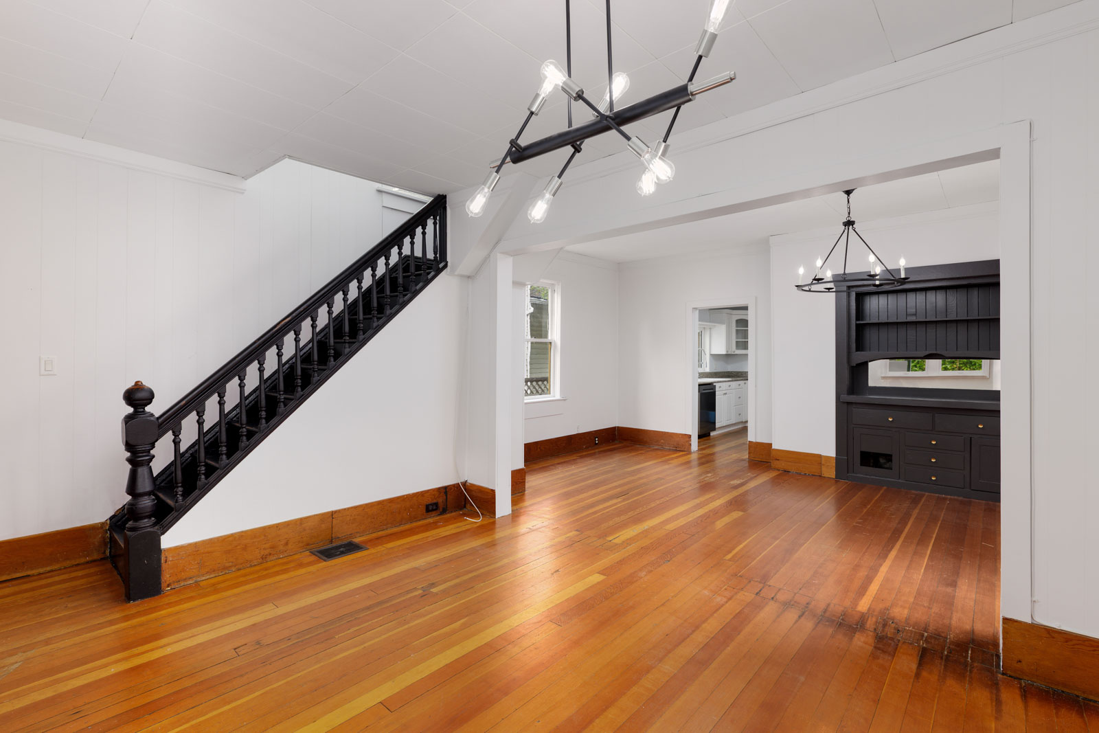 A spacious room with polished wood floors, white walls, a black staircase, modern chandelier, and a built-in black cabinet near an open kitchen doorway.