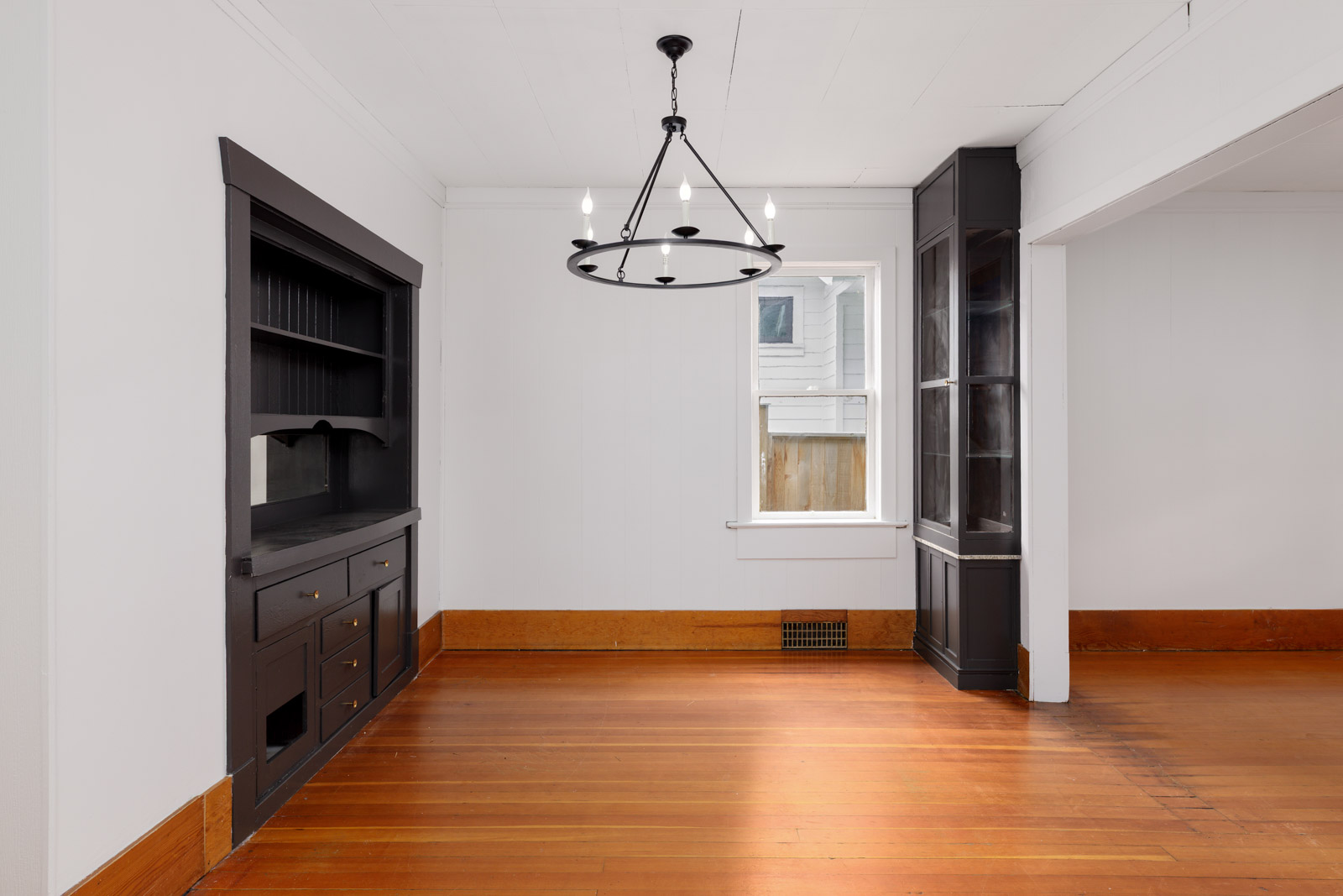 Unfurnished room with hardwood floors, white walls, black built-in cabinets, a round black chandelier, and a window letting in natural light.