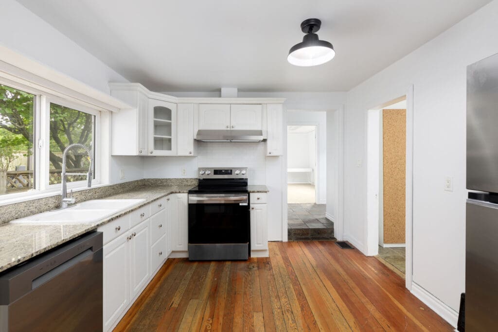 A kitchen with white cabinets, granite countertops, stainless steel appliances, a large window, and hardwood floors.