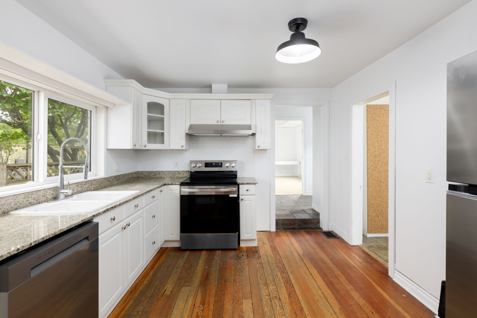 A kitchen with white cabinets, granite countertops, stainless steel appliances, a large window, and hardwood floors.
