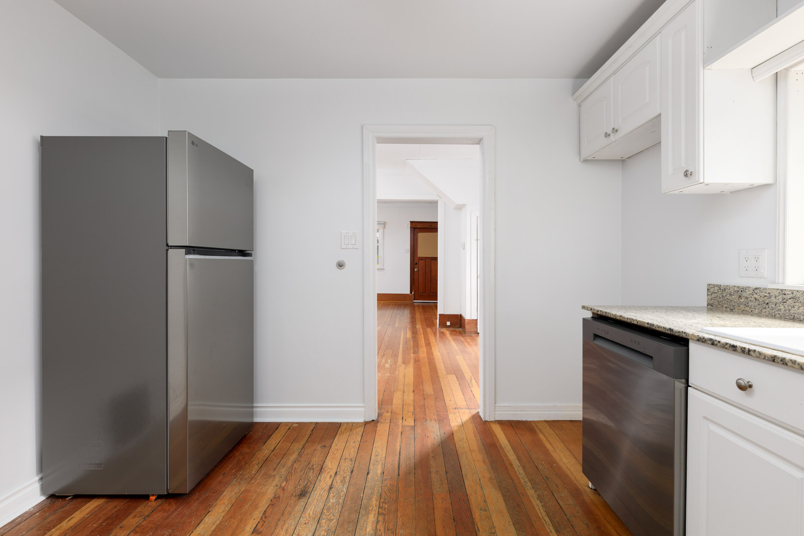 A kitchen with stainless steel appliances, white cabinets, and wood flooring, leading to a bright room with more wood floors.