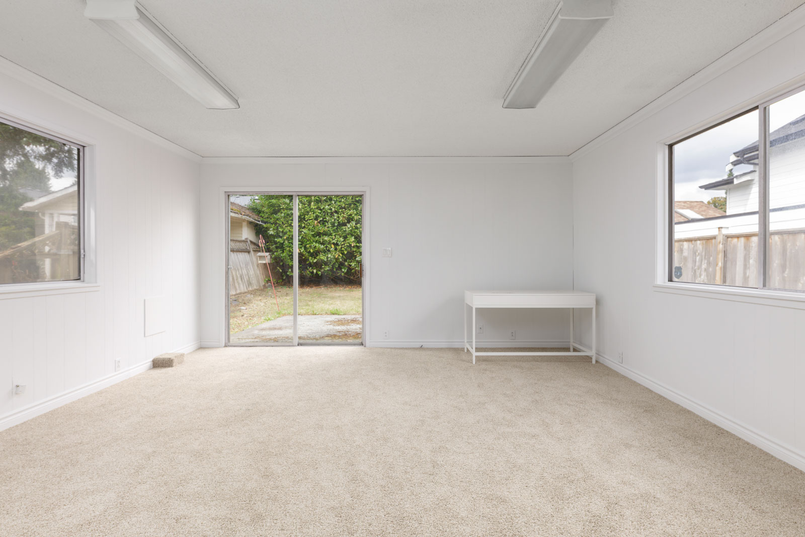An empty room with beige carpet, white walls, large windows, a glass sliding door, and a small white desk positioned near one window.