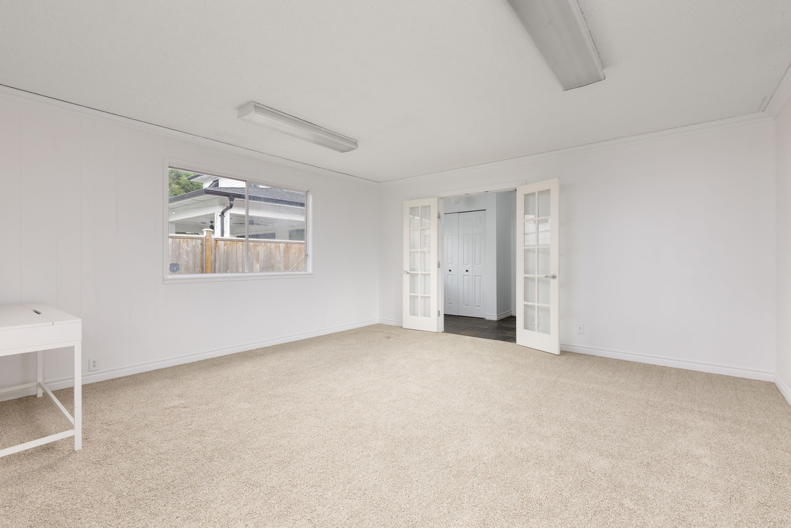 A bright, empty room with beige carpet, white walls, a window, fluorescent ceiling lights, and double glass doors leading to another area.