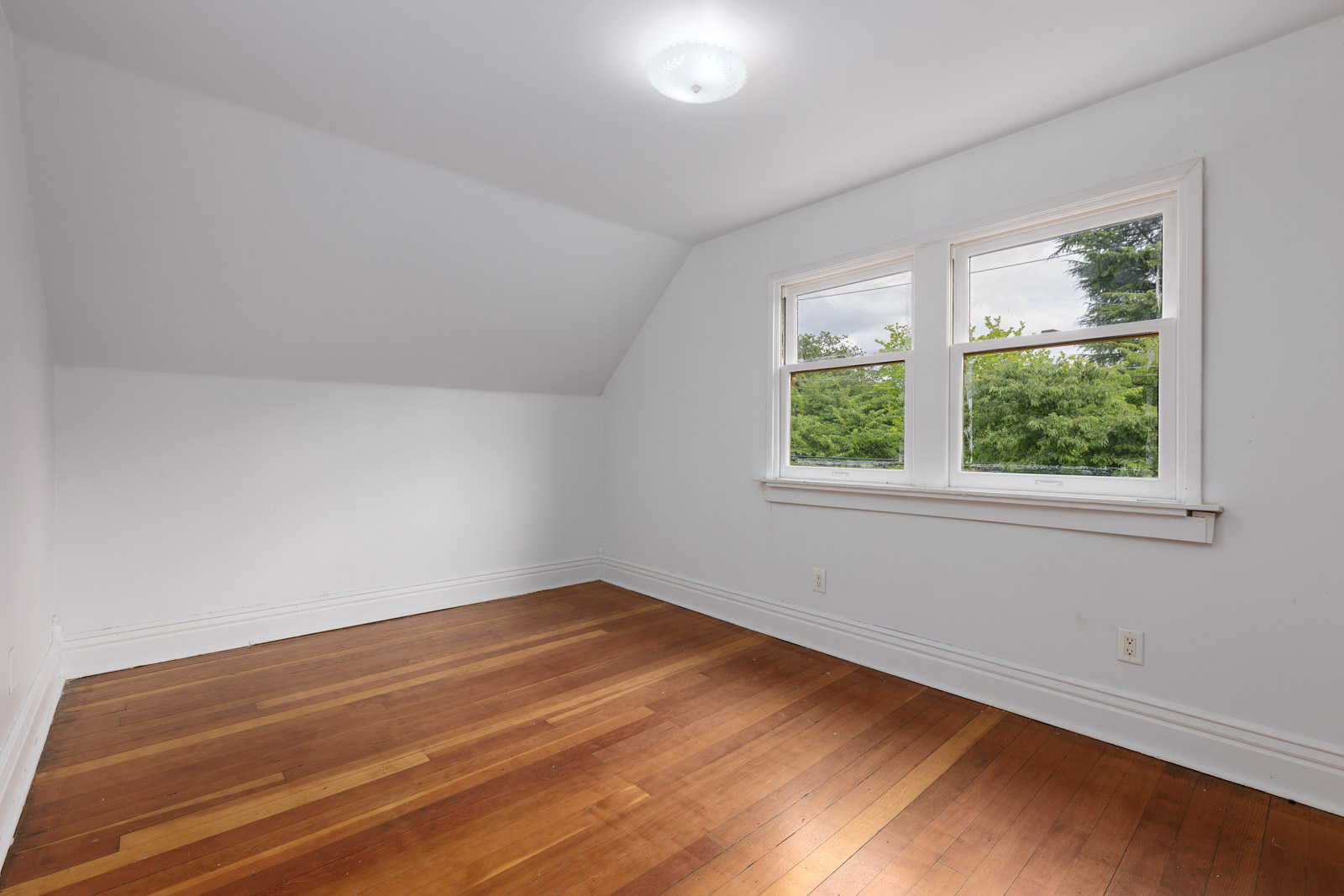 Unfurnished room with white walls, hardwood floor, sloped ceiling, ceiling light, and two windows overlooking greenery.