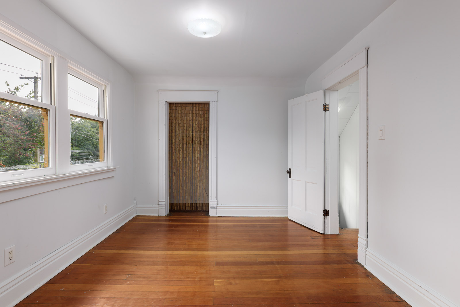 Empty room with white walls, hardwood floor, three windows, a ceiling light, an open door, and a recessed area with a brown curtain.