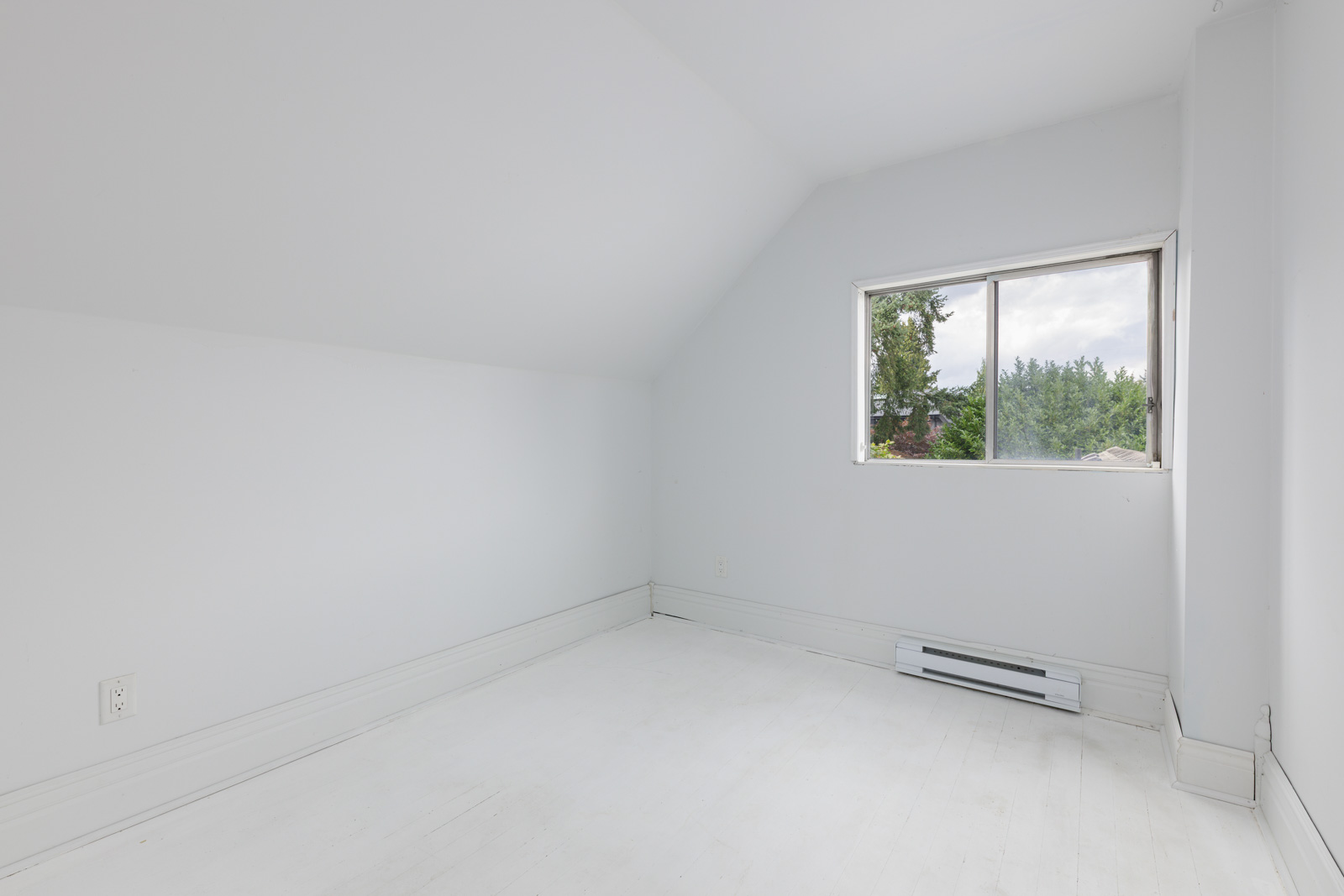 Empty white room with sloped ceiling, single window showing trees outside, white floor, wall heater, and electrical outlet visible.