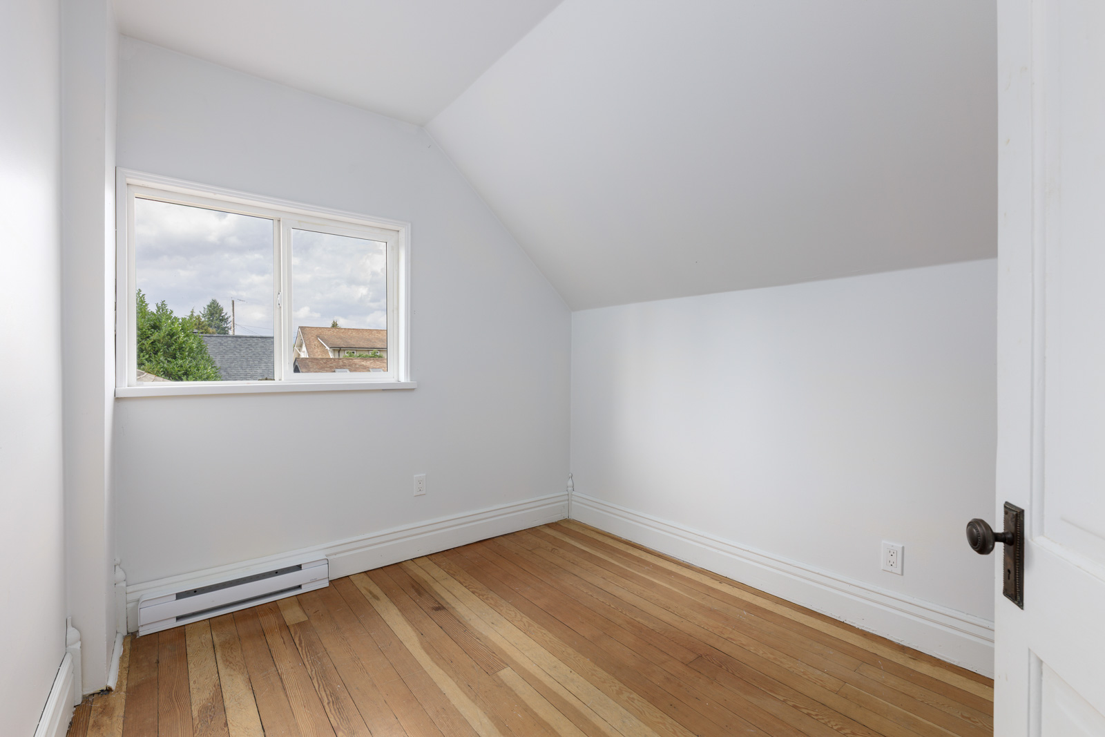 Small, empty room with white walls, sloped ceiling, hardwood floor, and a window showing a view of rooftops and trees outside.