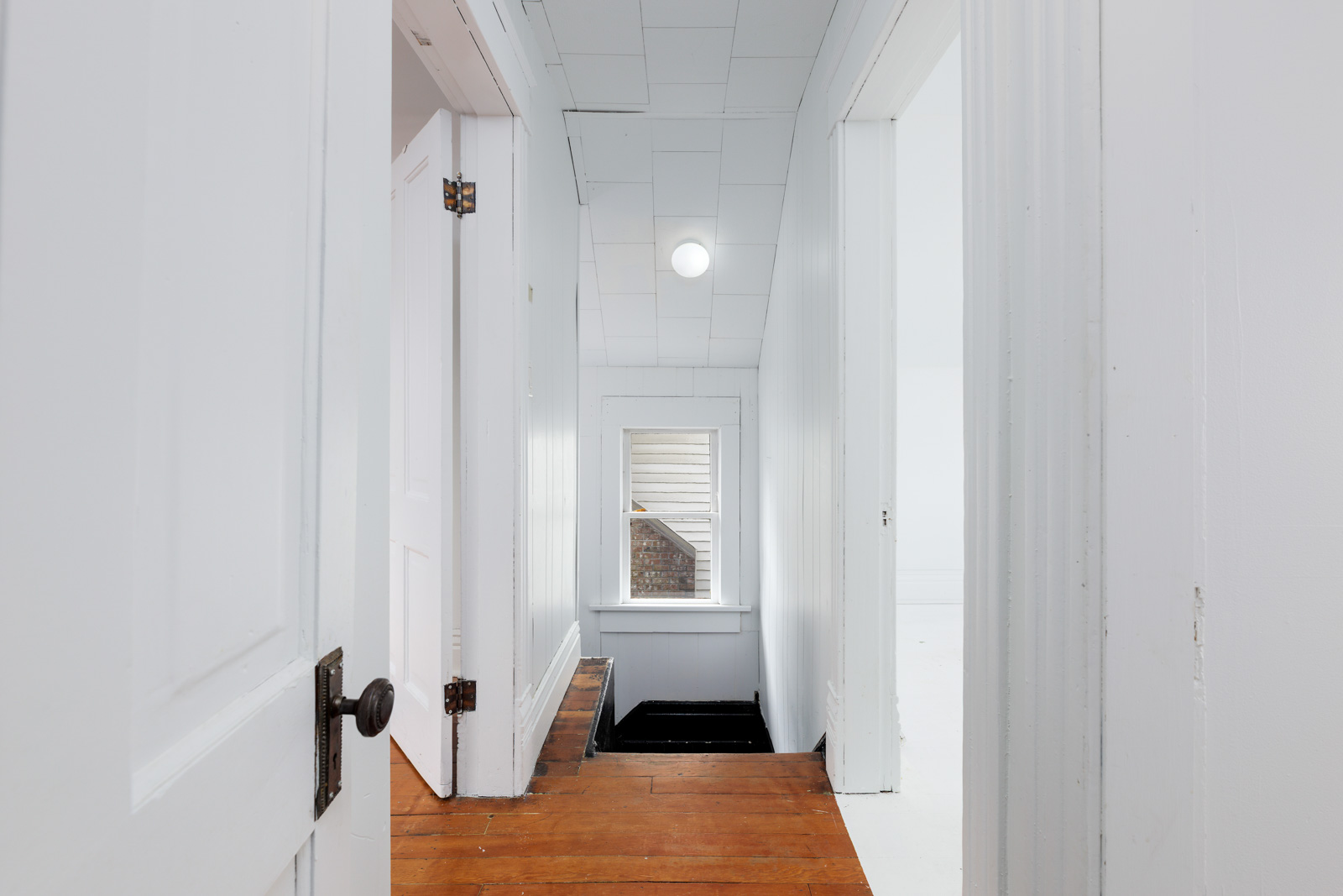 Narrow hallway with white walls and ceiling, wooden floor, open doorways, and a small staircase leading down near a window.