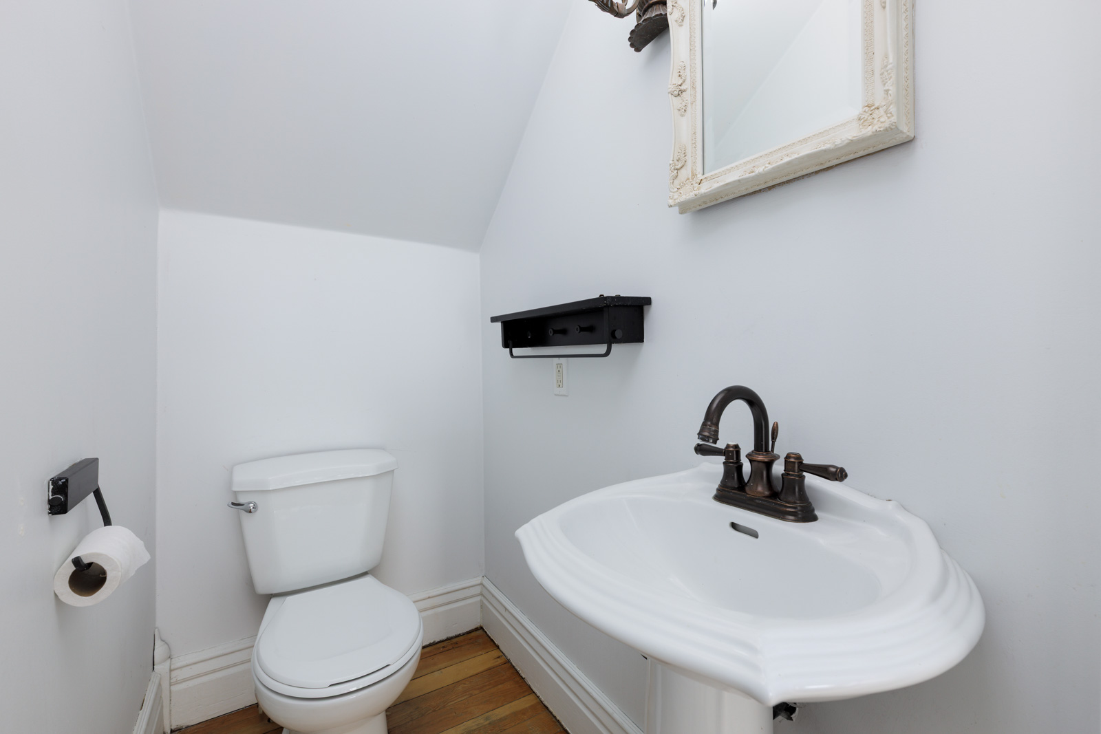 Small bathroom with a white toilet, pedestal sink with dark faucet, wall-mounted mirror, toilet paper holder, and wooden floor.