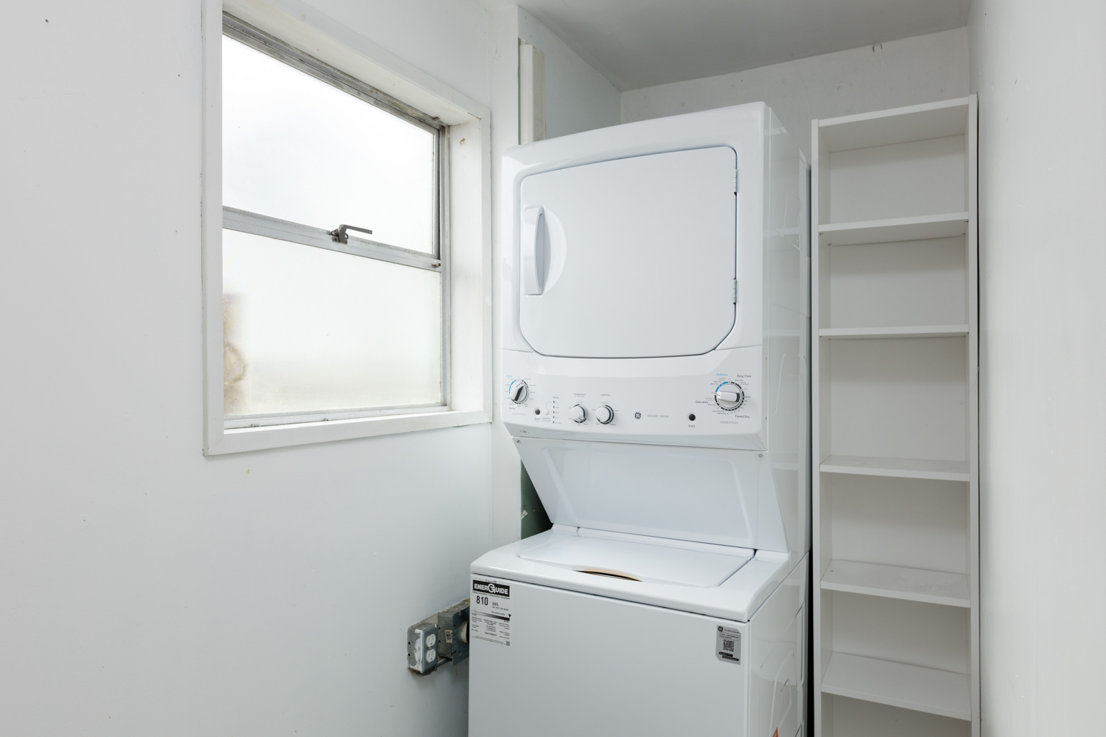 A stacked washer and dryer unit next to an empty white shelving unit in a small laundry room with a window.