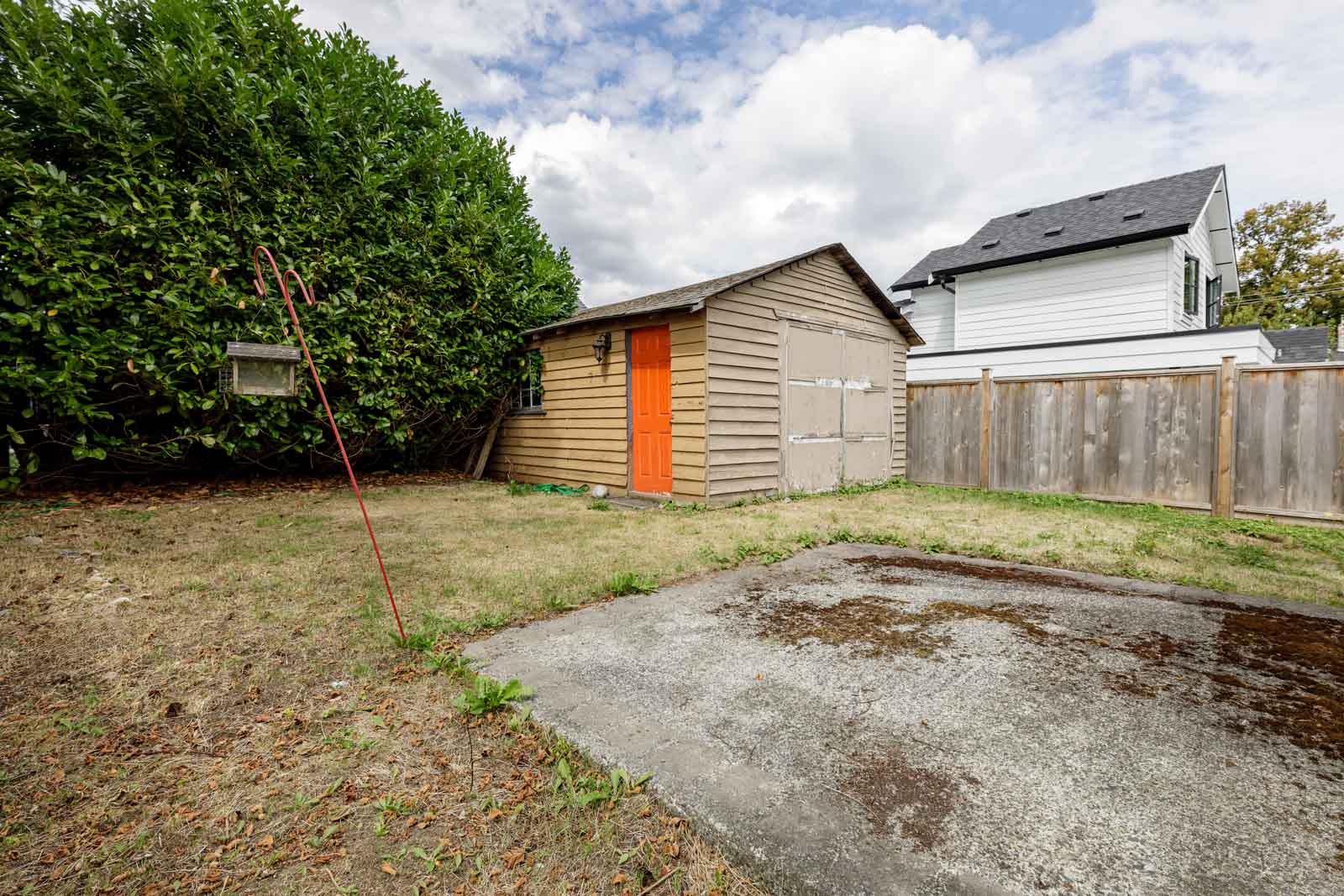 A small wooden shed with orange doors sits in a fenced backyard with patchy grass, a concrete slab, and a bush on the left under a partly cloudy sky.