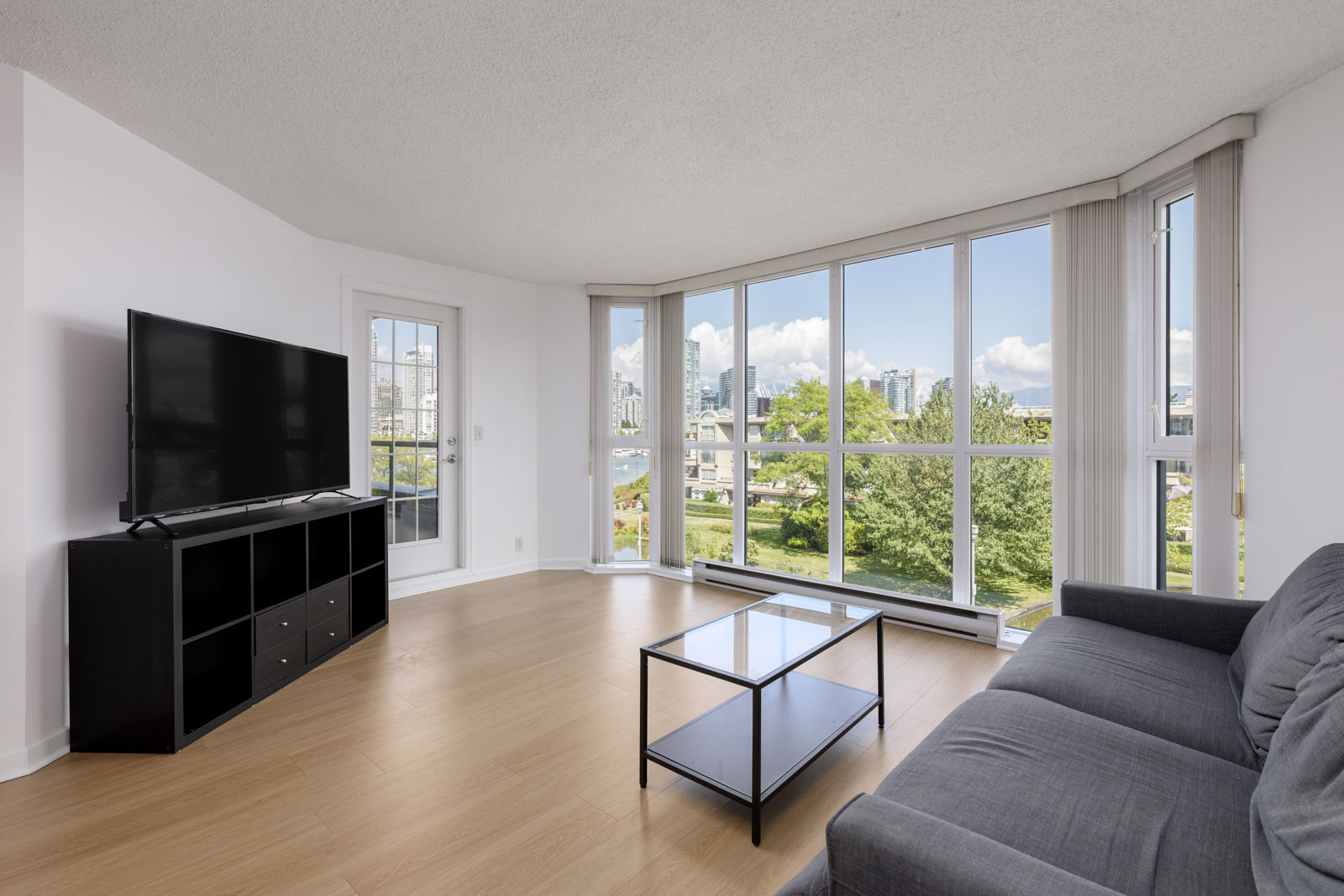 Bright living room with large corner windows, a gray sofa, black TV stand with a flat-screen TV, glass coffee table, and laminate wood flooring. City and greenery visible outside.