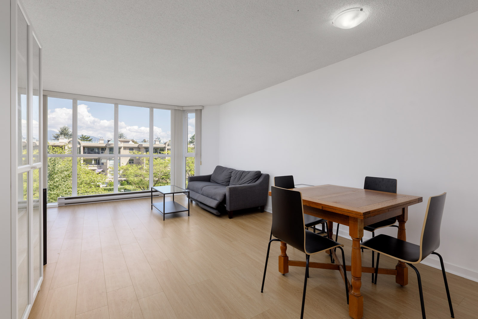 A bright living and dining area with wood flooring, a gray sofa, a small table, four black chairs, and large windows overlooking trees and buildings.