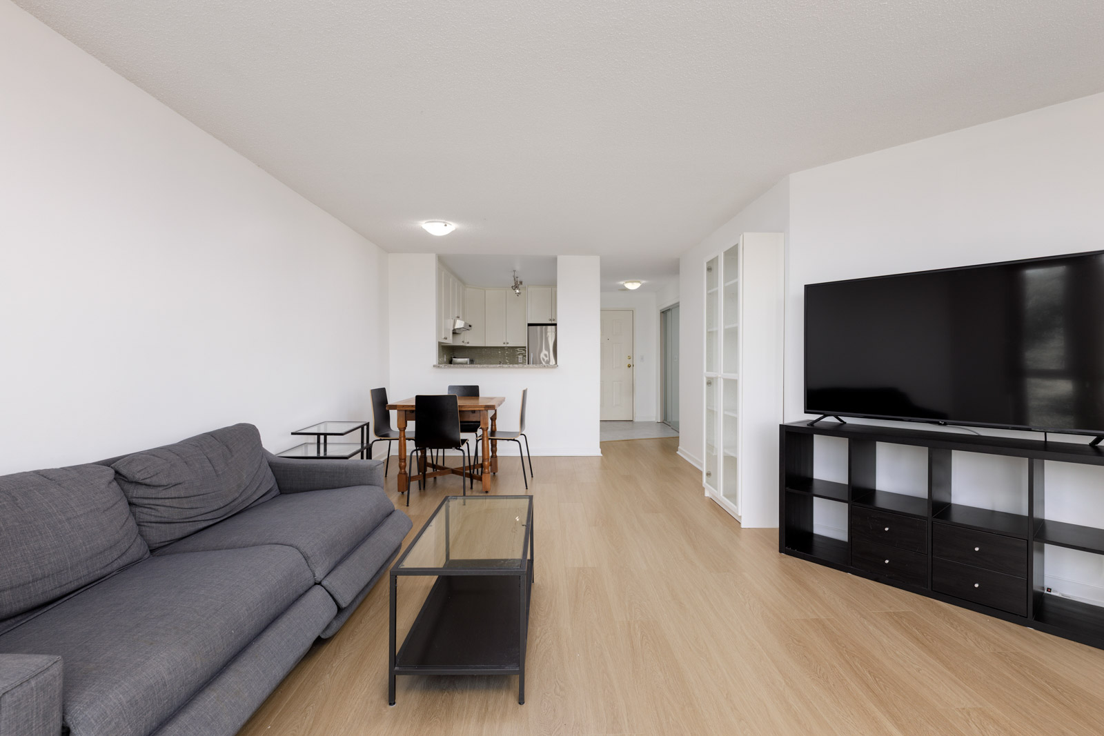 A modern living room with a gray sofa, glass coffee table, dining area, flat-screen TV on a black stand, and a view of the kitchen in the background.