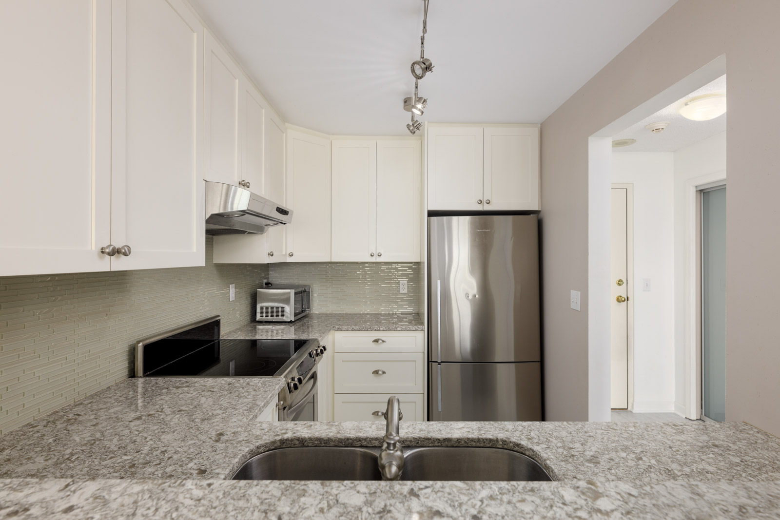 Modern kitchen with white cabinets, stainless steel appliances, granite countertops, and a mosaic tile backsplash, viewed from behind a sink island.