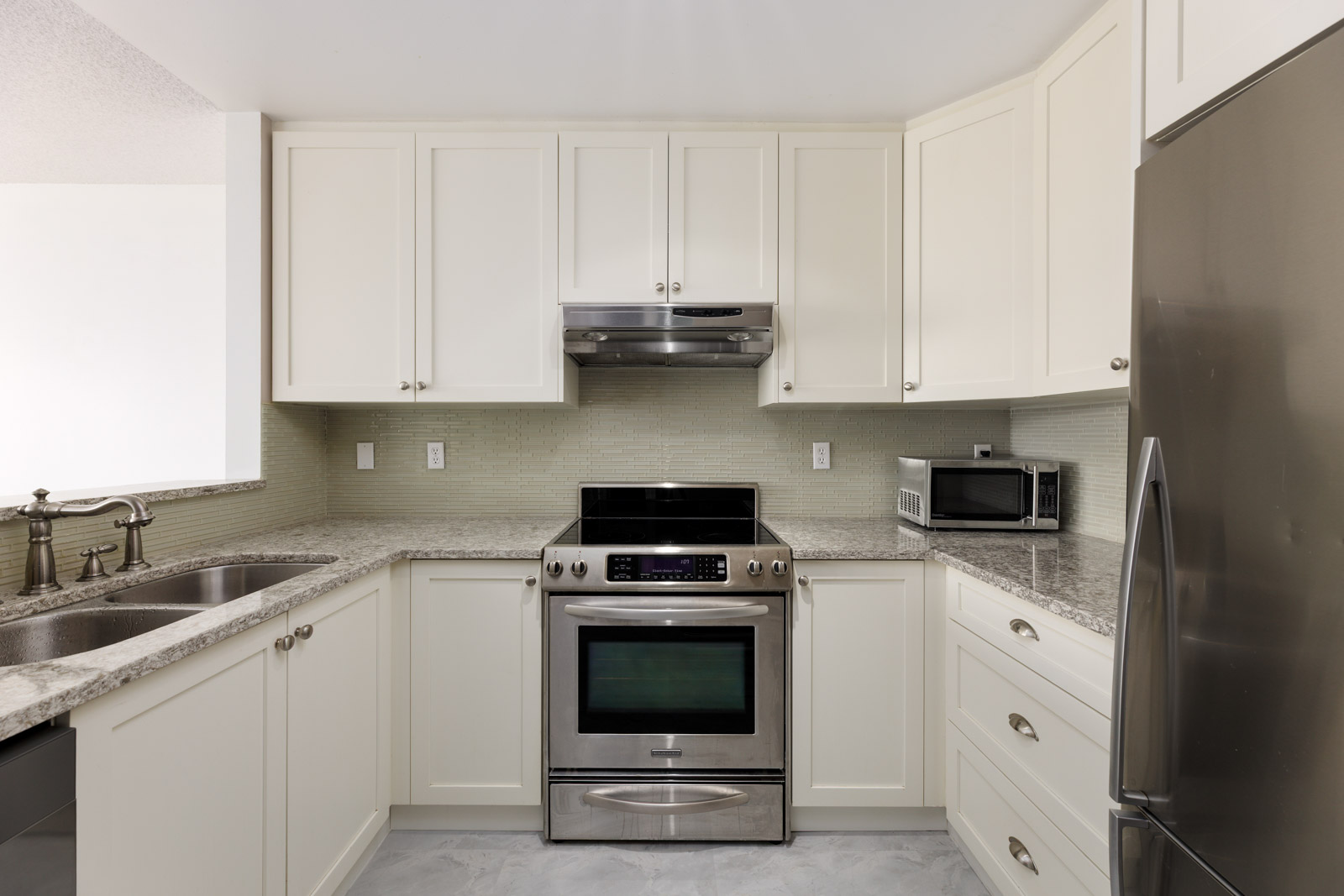 Modern kitchen with white cabinets, stainless steel appliances, electric stove, double sink, small microwave, and light-colored countertop and backsplash.