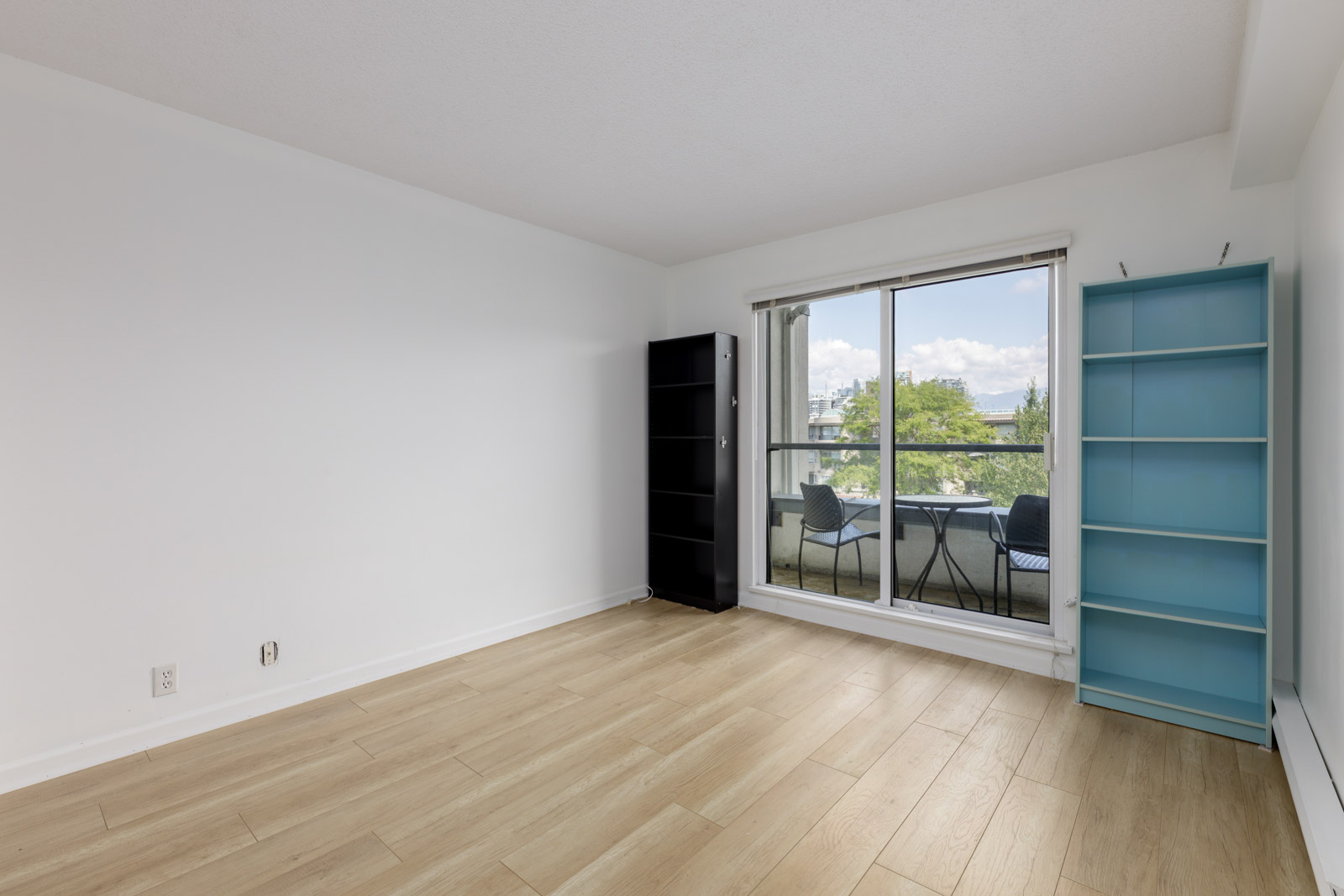An empty room with light wood flooring, white walls, a black shelf and a blue shelf, with sliding glass doors leading to a balcony with outdoor chairs and a table.