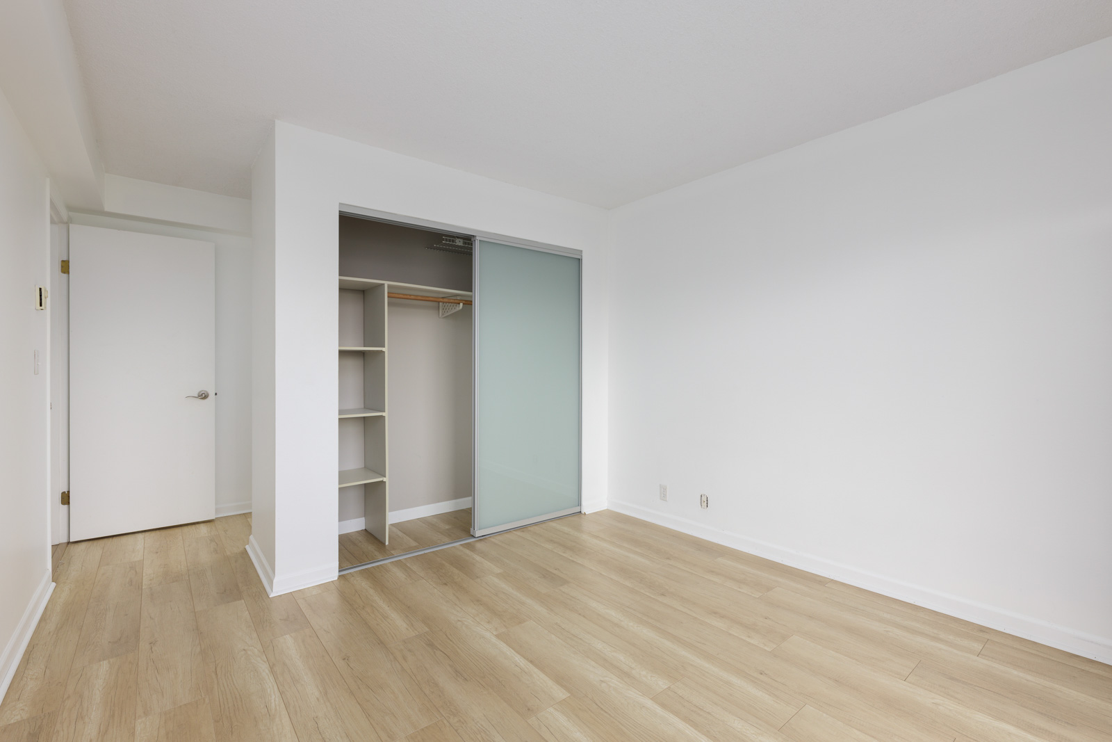 Empty room with light wood flooring, white walls, a closet with sliding doors, and a closed white door on the left.