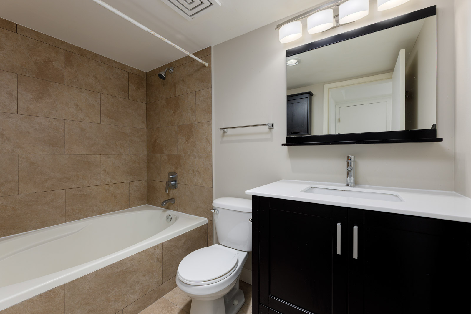 Modern bathroom with a tiled bathtub-shower combo, white toilet, dark vanity with sink, large mirror, and overhead lighting.