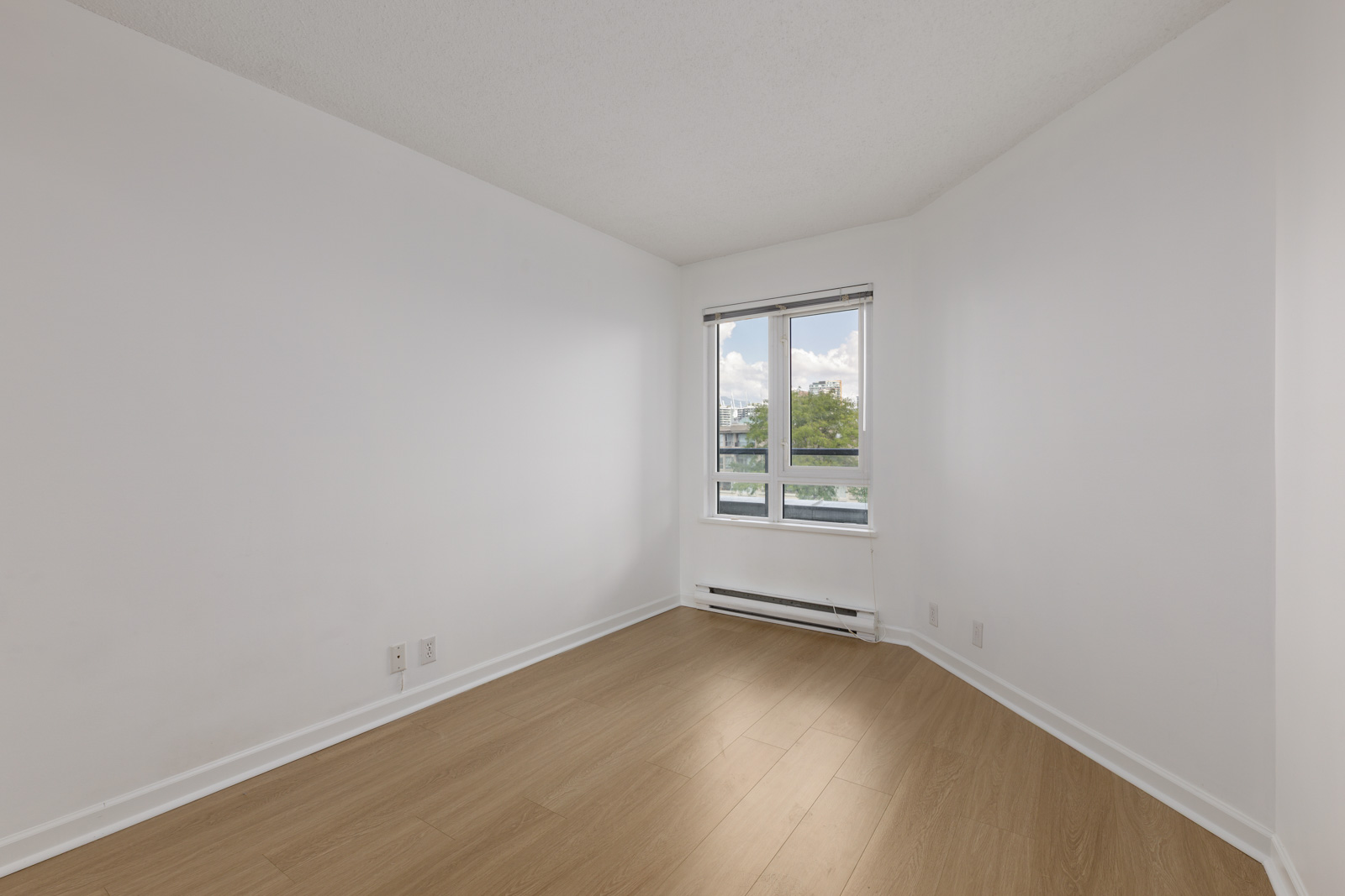 Empty room with white walls, a large window letting in natural light, light wood flooring, baseboard heater, and electrical outlets visible.