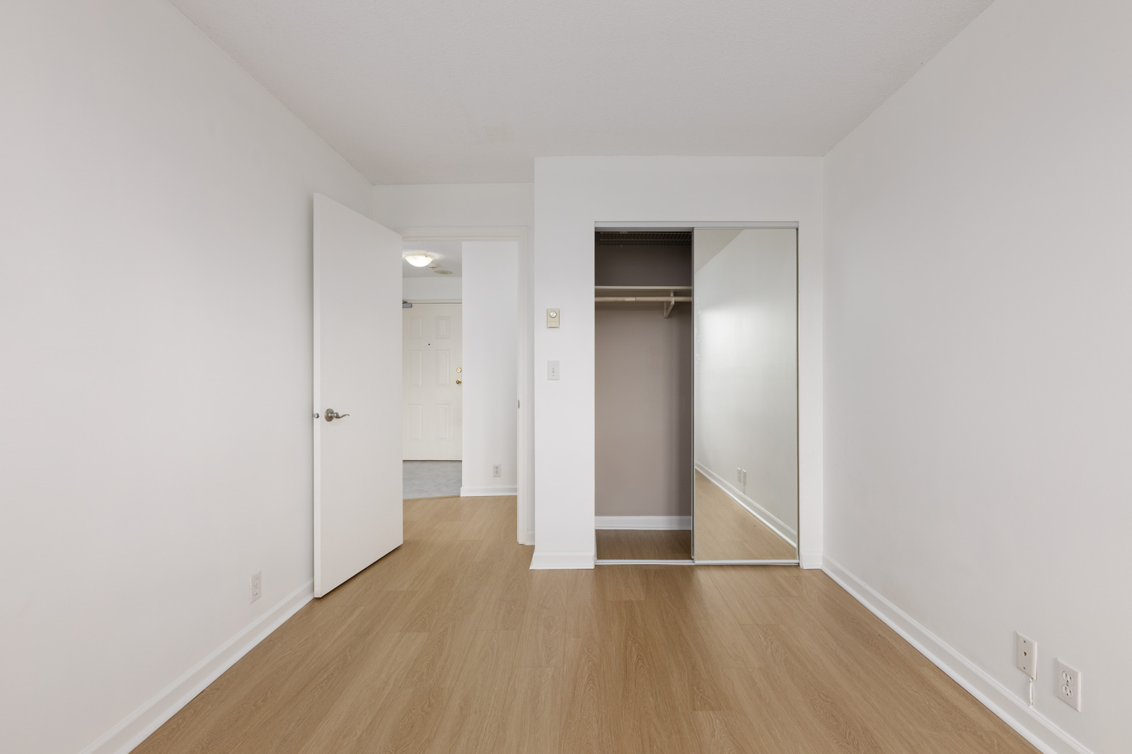 Empty room with white walls, light wood flooring, an open door, and a closet with sliding mirrored doors.