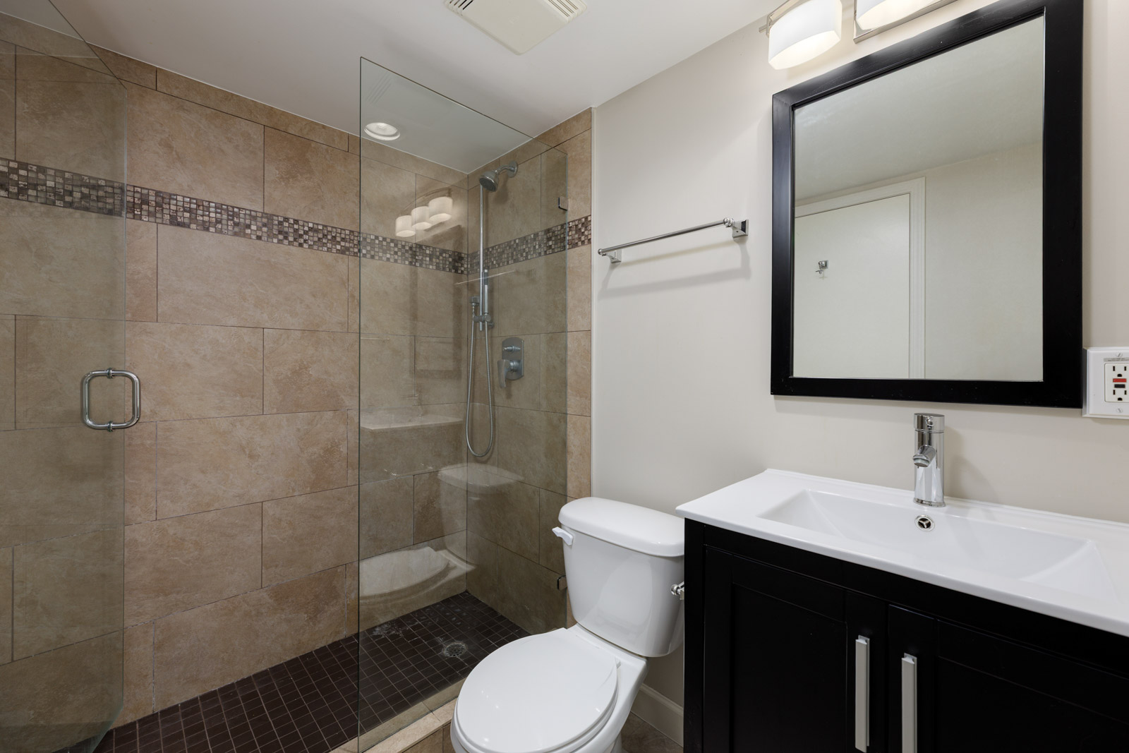 Modern bathroom with glass-enclosed shower, beige tile walls, white toilet, and black vanity with sink and mirror.