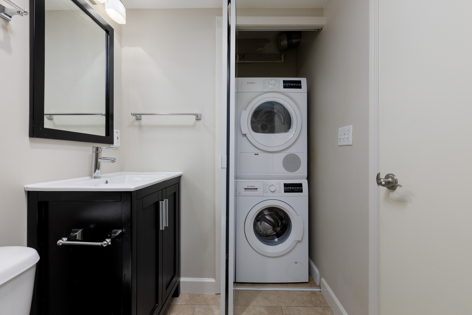 A bathroom with a black vanity, white sink, wall mirror, and towel rack, next to a closet containing stacked washer and dryer units.