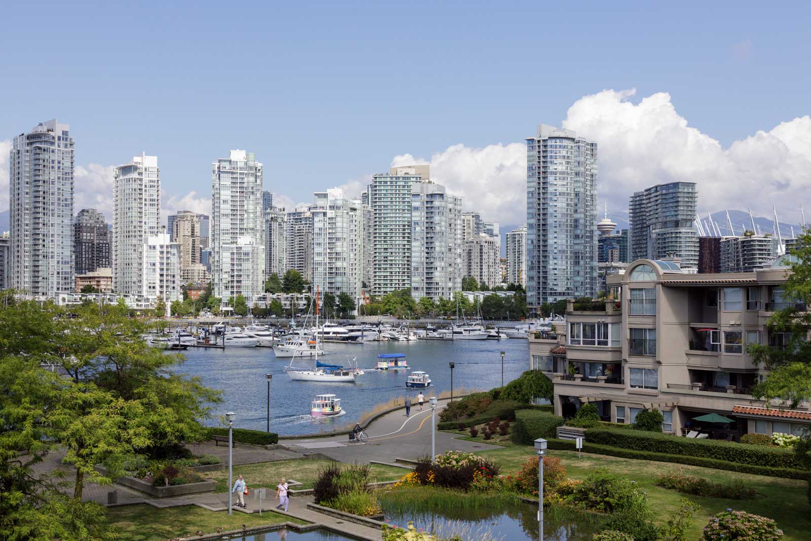 High-rise buildings and waterfront condos surround a marina with boats docked and sailing, set against a partly cloudy sky in an urban cityscape.