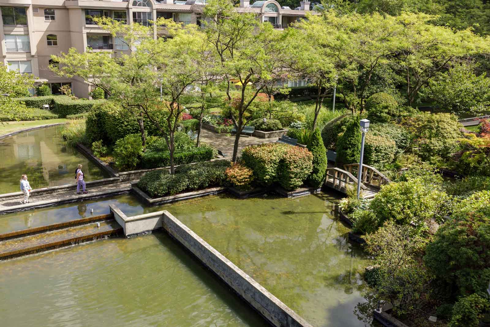 An apartment complex courtyard with landscaped gardens, walking paths, small bridges, and shallow ponds; two people walk along the paved area.