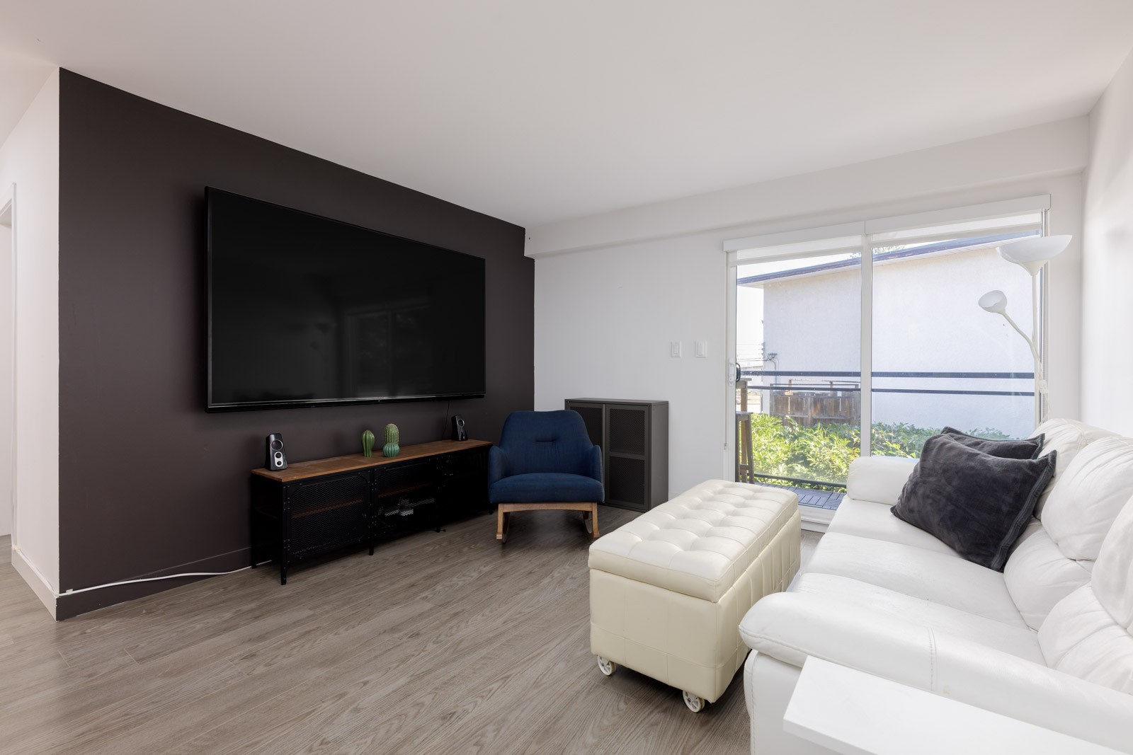 A modern living room with a large wall-mounted TV, a black entertainment unit, a blue chair, a white sofa with black pillows, a white ottoman, and a floor lamp by a window.