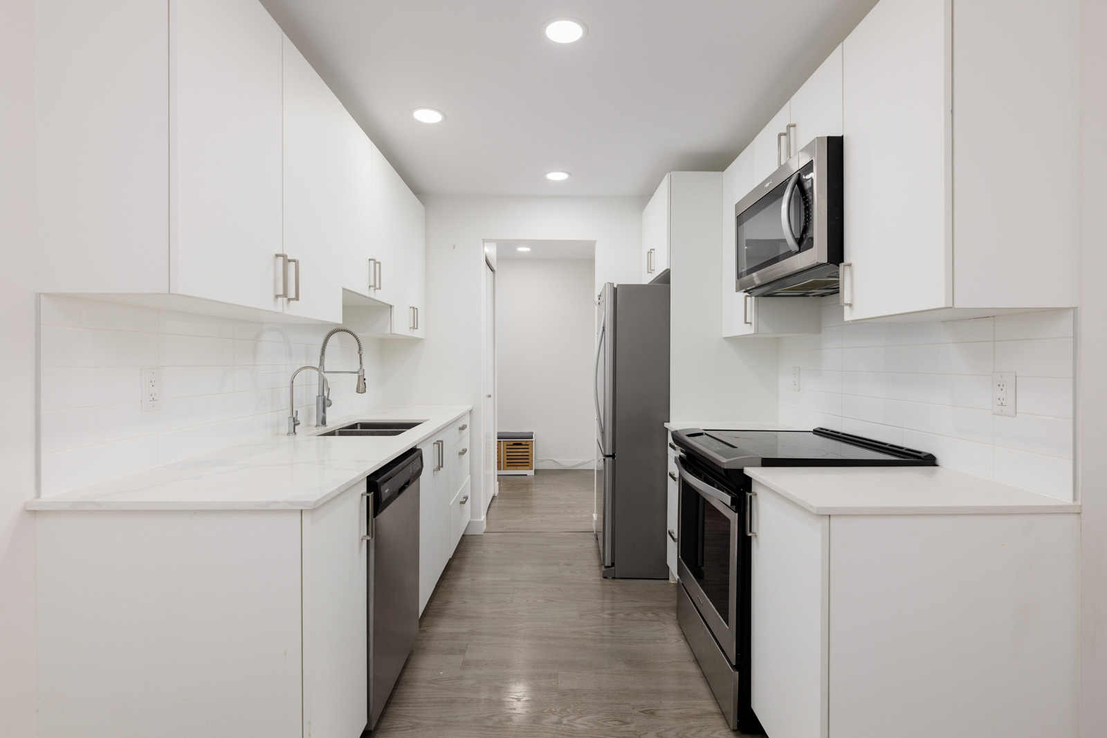Modern galley kitchen with white cabinets, stainless steel appliances, electric stove, built-in microwave, double sink, and light wood flooring under recessed ceiling lights.