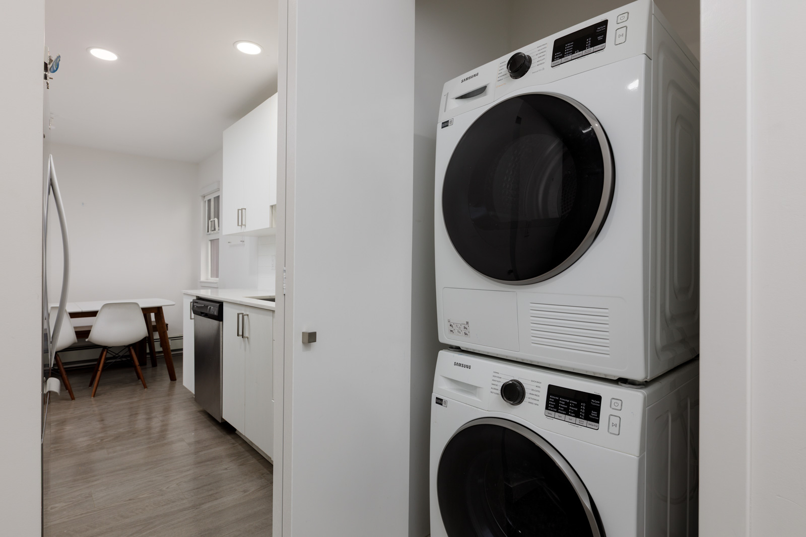 A stacked washer and dryer are installed in a small laundry nook next to a modern kitchen with white cabinets and a dining area visible in the background.