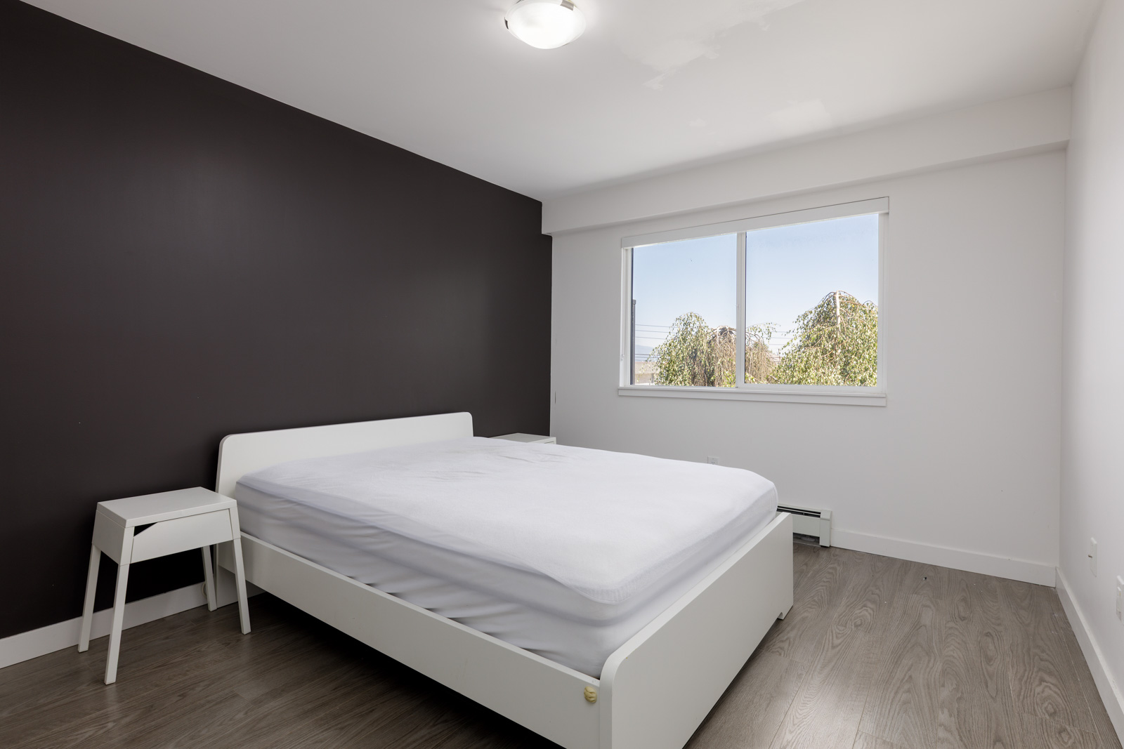 Minimalist bedroom with a white bed, small side table, dark accent wall, large window, and wood flooring.