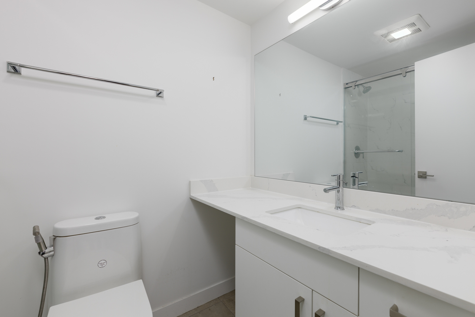 Modern bathroom with white walls, a large mirror above a white vanity, a sink, a toilet, and a glass-enclosed shower in the background.