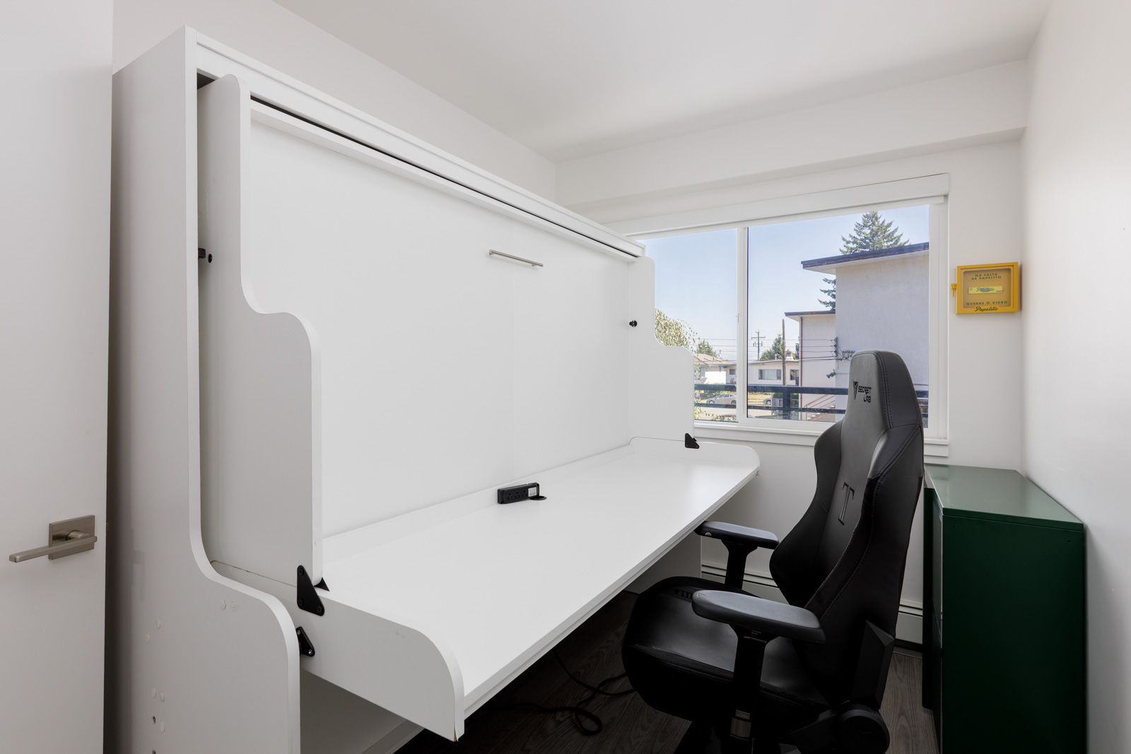 A compact office setup with a fold-down white desk, black office chair, green cabinet, and a window letting in natural light.